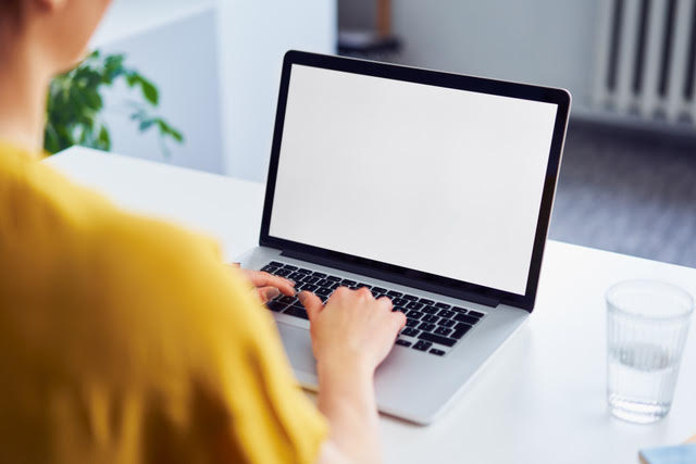 Person using a laptop with a blank white screen on a white desk, with a glass of water nearby.