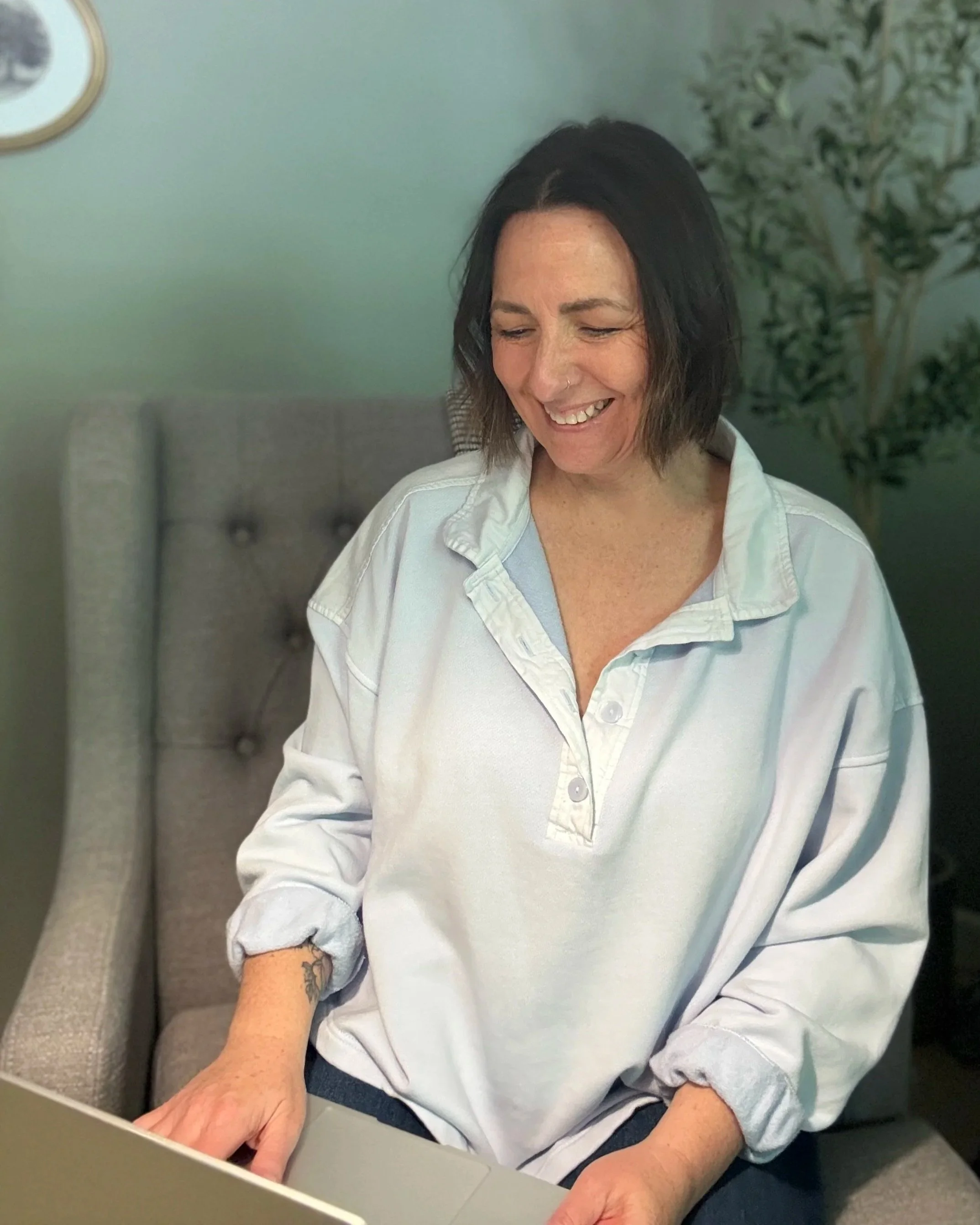 A woman with short dark hair and a nose piercing smiling while sitting in front of a gray upholstered headboard and a potted plant, wearing a white button-up shirt with rolled-up sleeves and jeans.