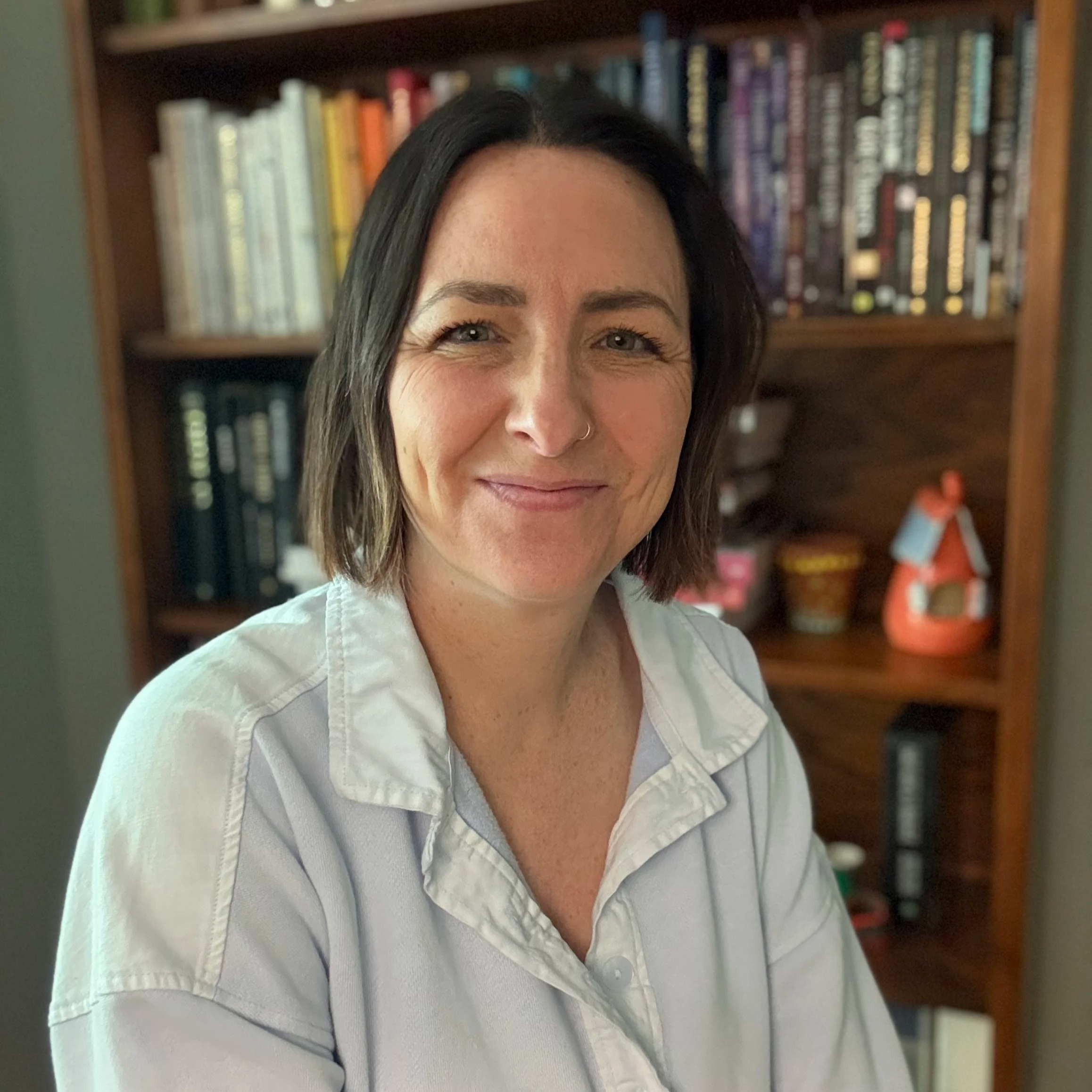 A woman with shoulder-length brown hair smiling, wearing a white shirt, standing in front of a bookshelf filled with books.