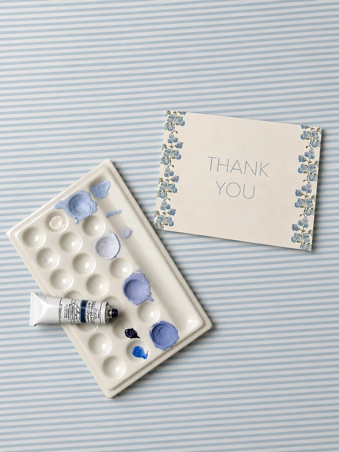 Palette with blue paint and a thank you card with floral border on striped background.