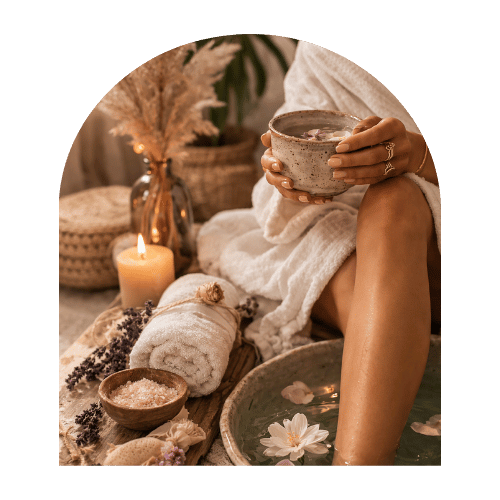 A person sitting with legs in a foot bath, holding a ceramic mug, surrounded by candles, towels, and spa decor.