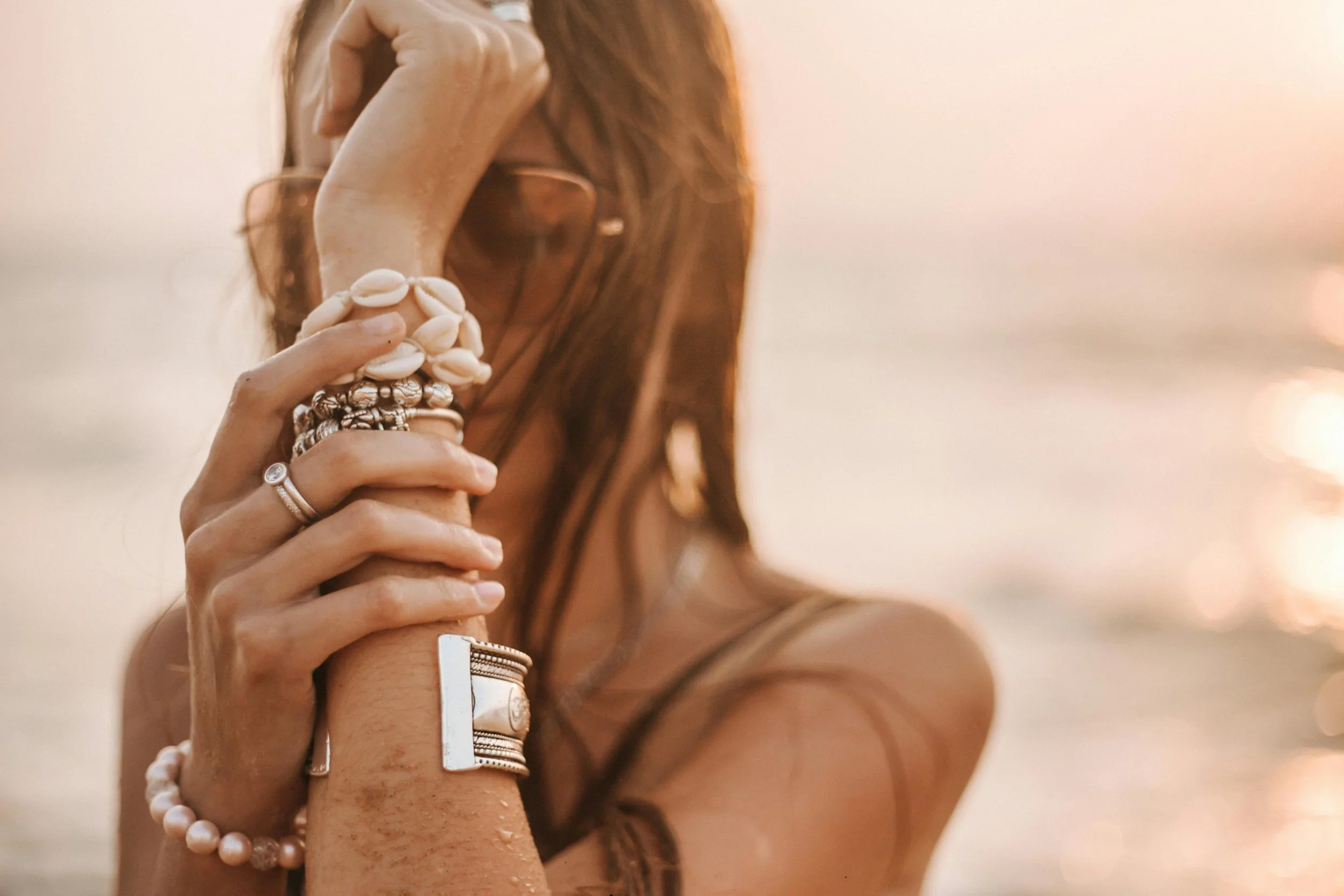 A woman wearing multiple rings and a pearl bracelet holding a mug with shells on it, near the water at sunset.
