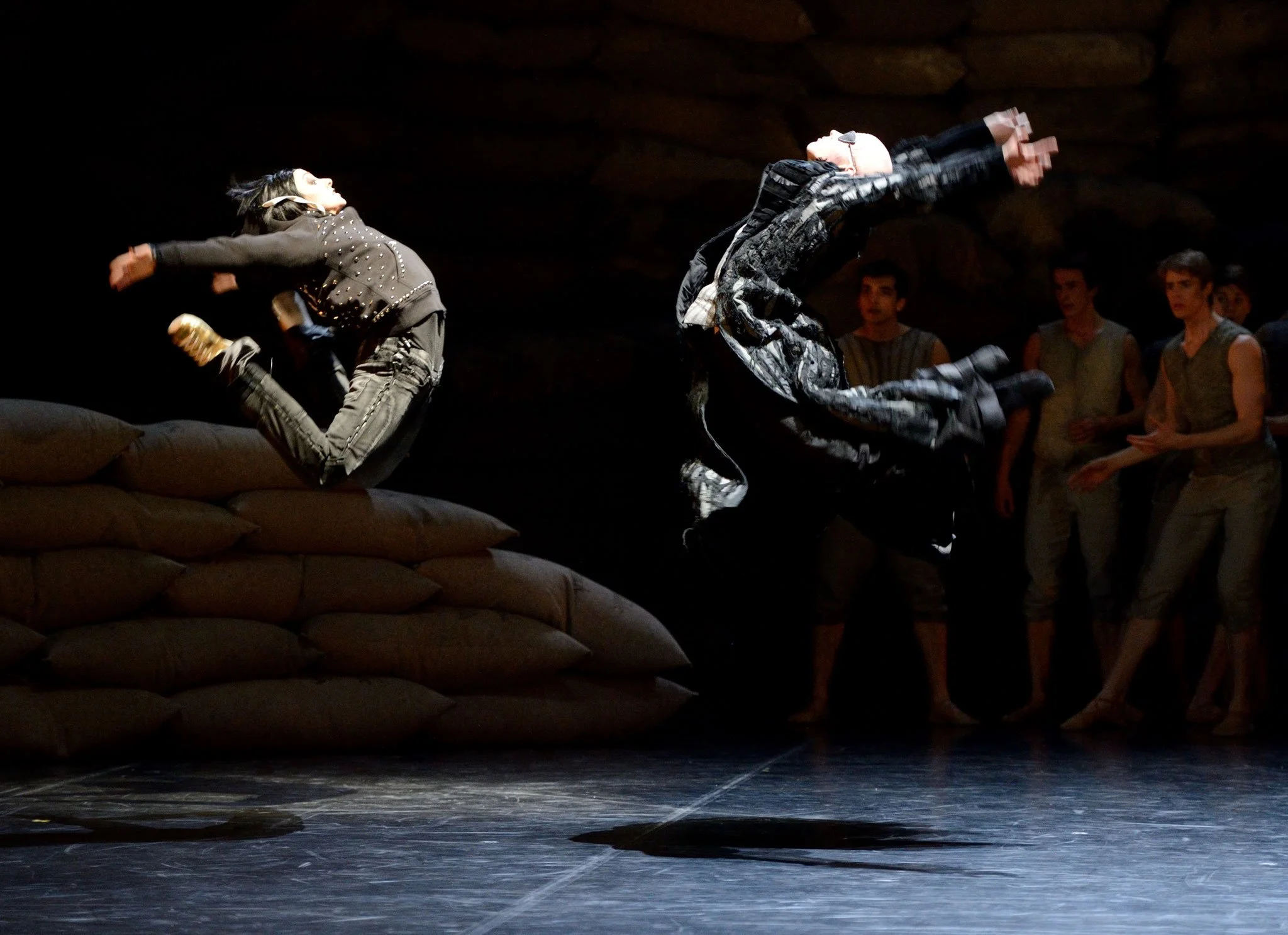 Two performers are mid-air in a dance or acrobatic move on a stage, with a group of people observing in the background.