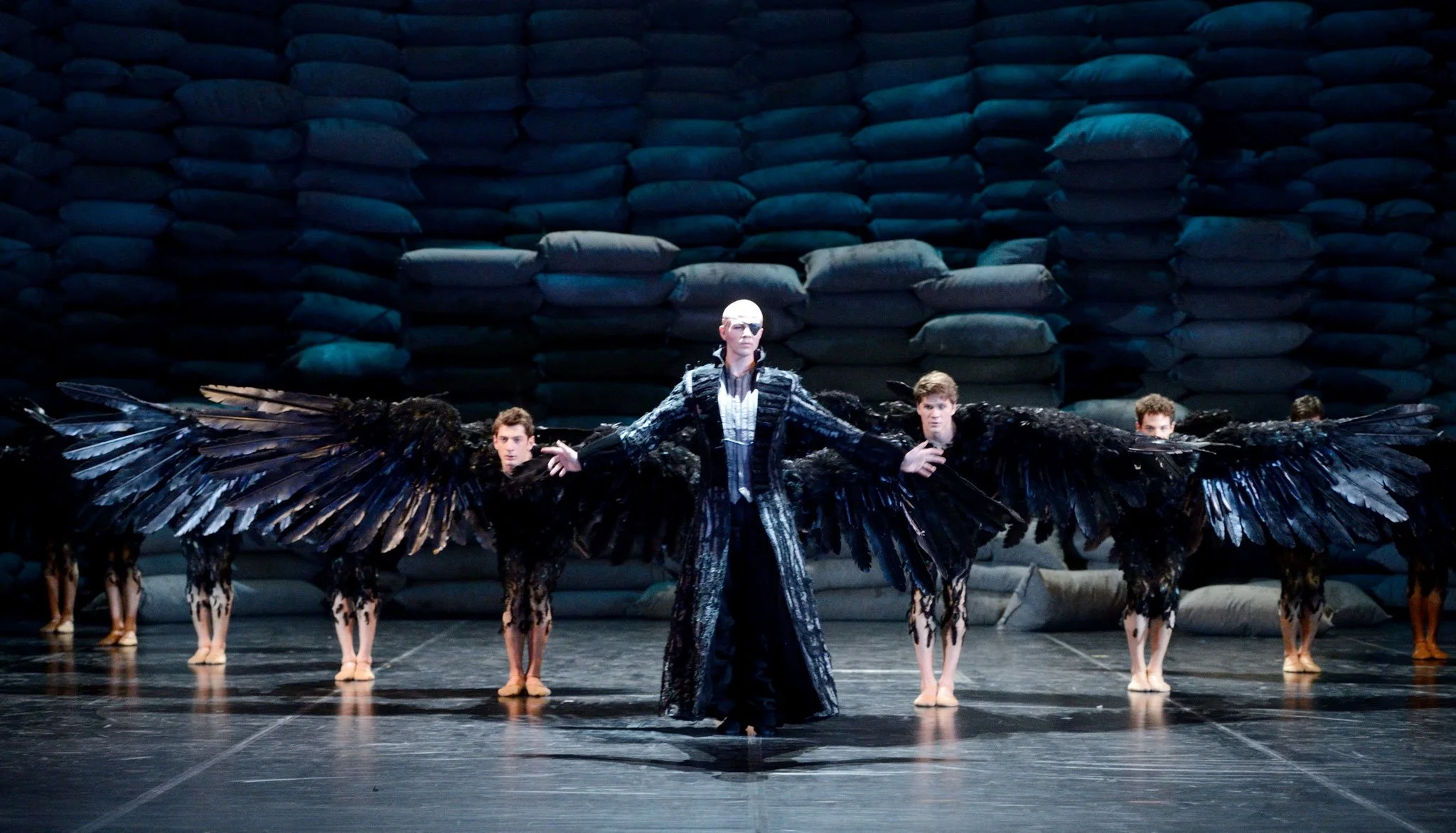 A theatrical performance featuring a central figure with a bald head and dark, metallic costume with large black wings, surrounded by dancers with similar wings, against a backdrop of stacked sacks.