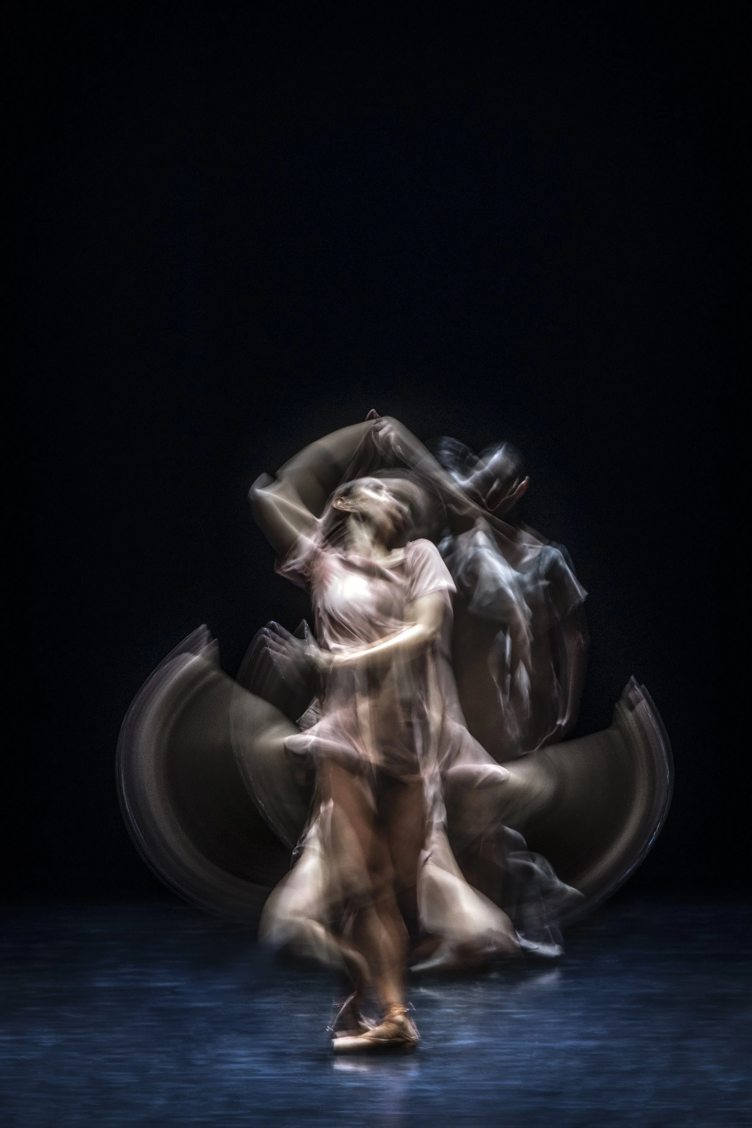 A dancer in a light-colored dress performs a movement on stage, with a dark background and motion blur creating a dynamic and abstract effect.