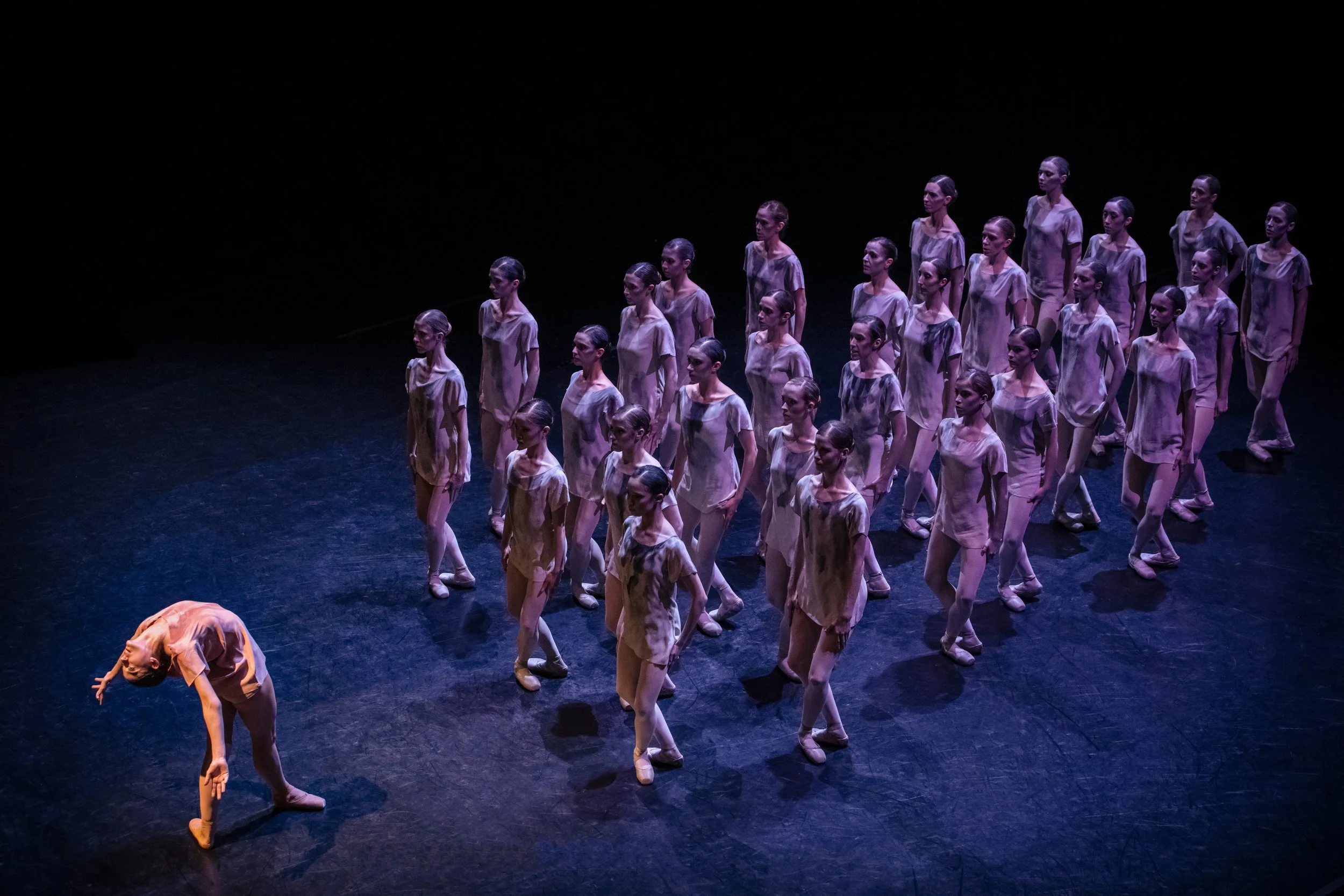 A group of young dancers performing on stage, with one dancer bending backwards and extending her arms, while the others stand in formation behind her, all dressed in matching costumes under stage lighting.