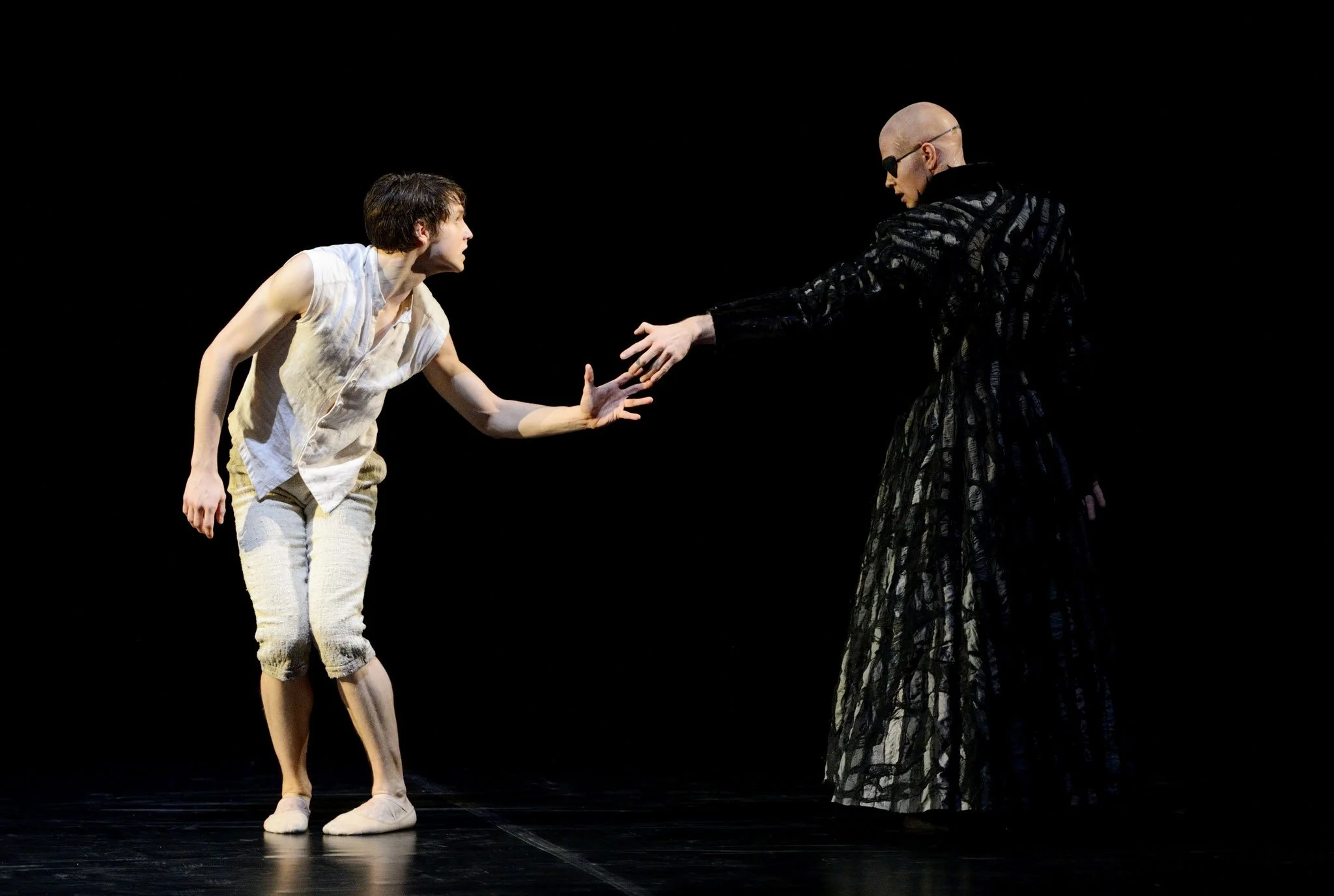 Two performers on stage, one dressed in light clothing with short dark hair, reaching out to a taller person with a bald head, and a dark patterned outfit, both engaging in a theatrical scene.