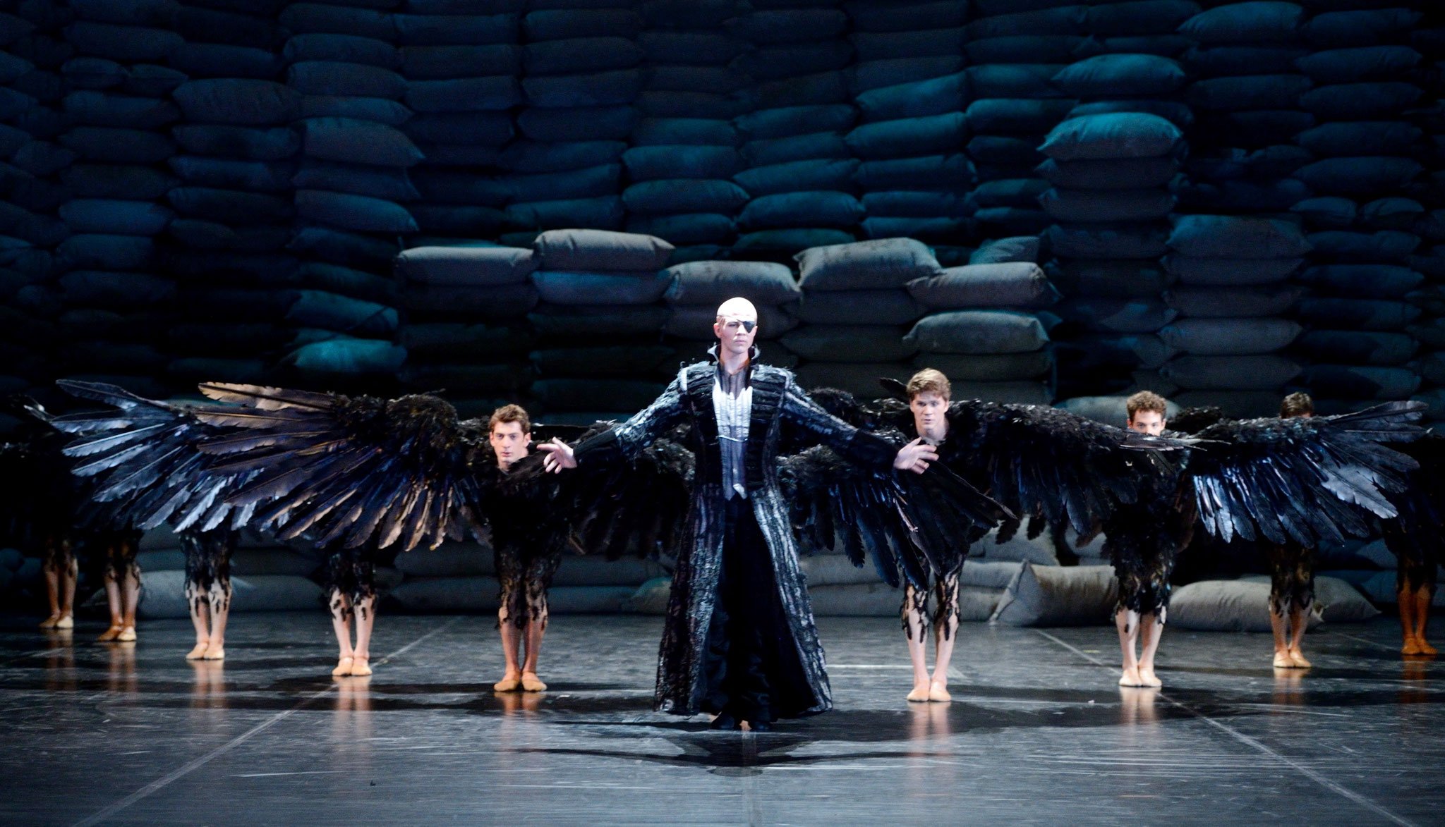 The image shows a stage performance with a central person dressed in dark, feathered costume, with large black wings. Surrounding them are dancers in similar feathered costumes, standing in a formation. The background features a wall made of stacked 
