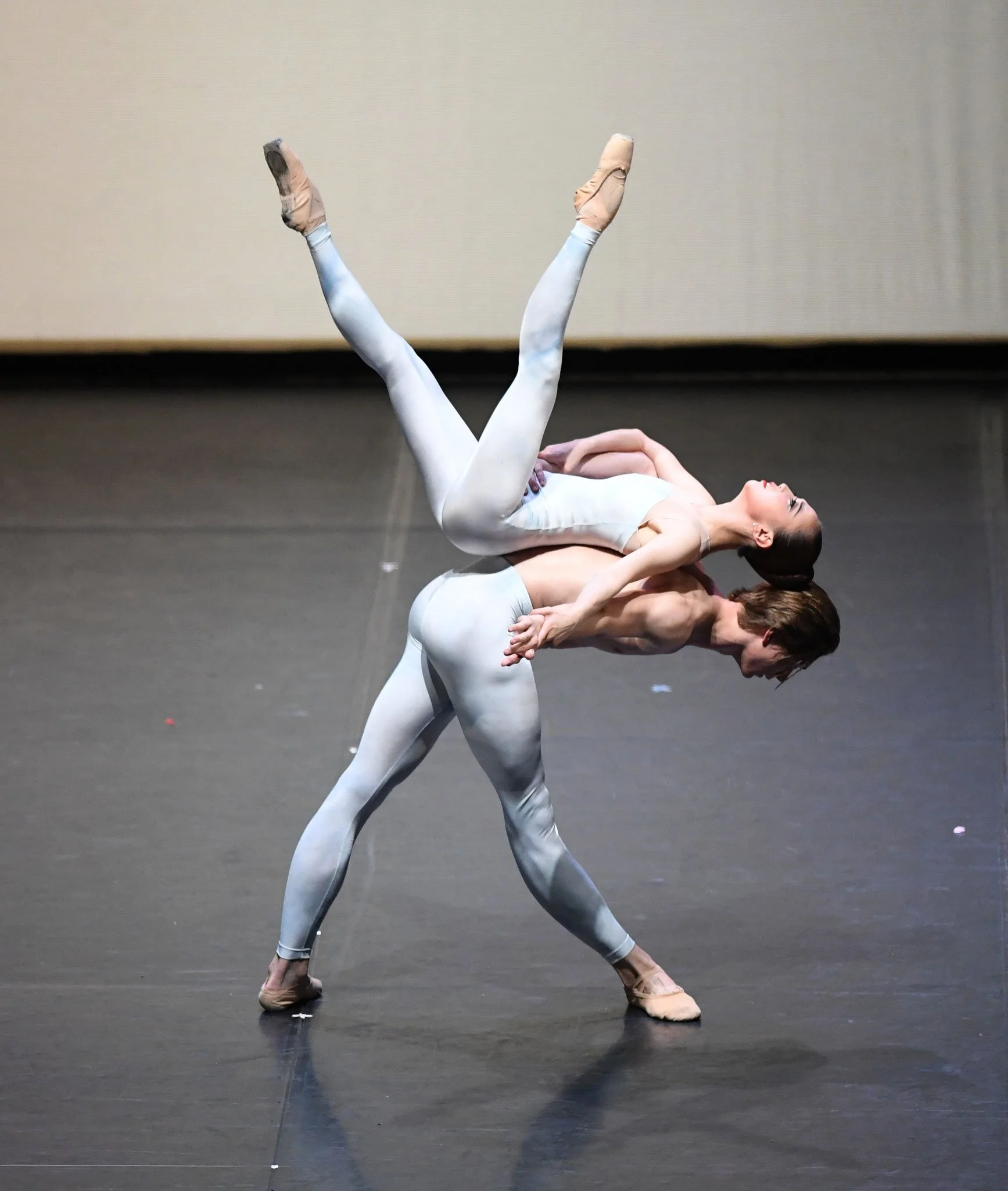 Two ballet dancers performing on stage in white costumes: one dancer is balanced on the other's back with legs extended upward, while the second dancer supports with arms and balance. 