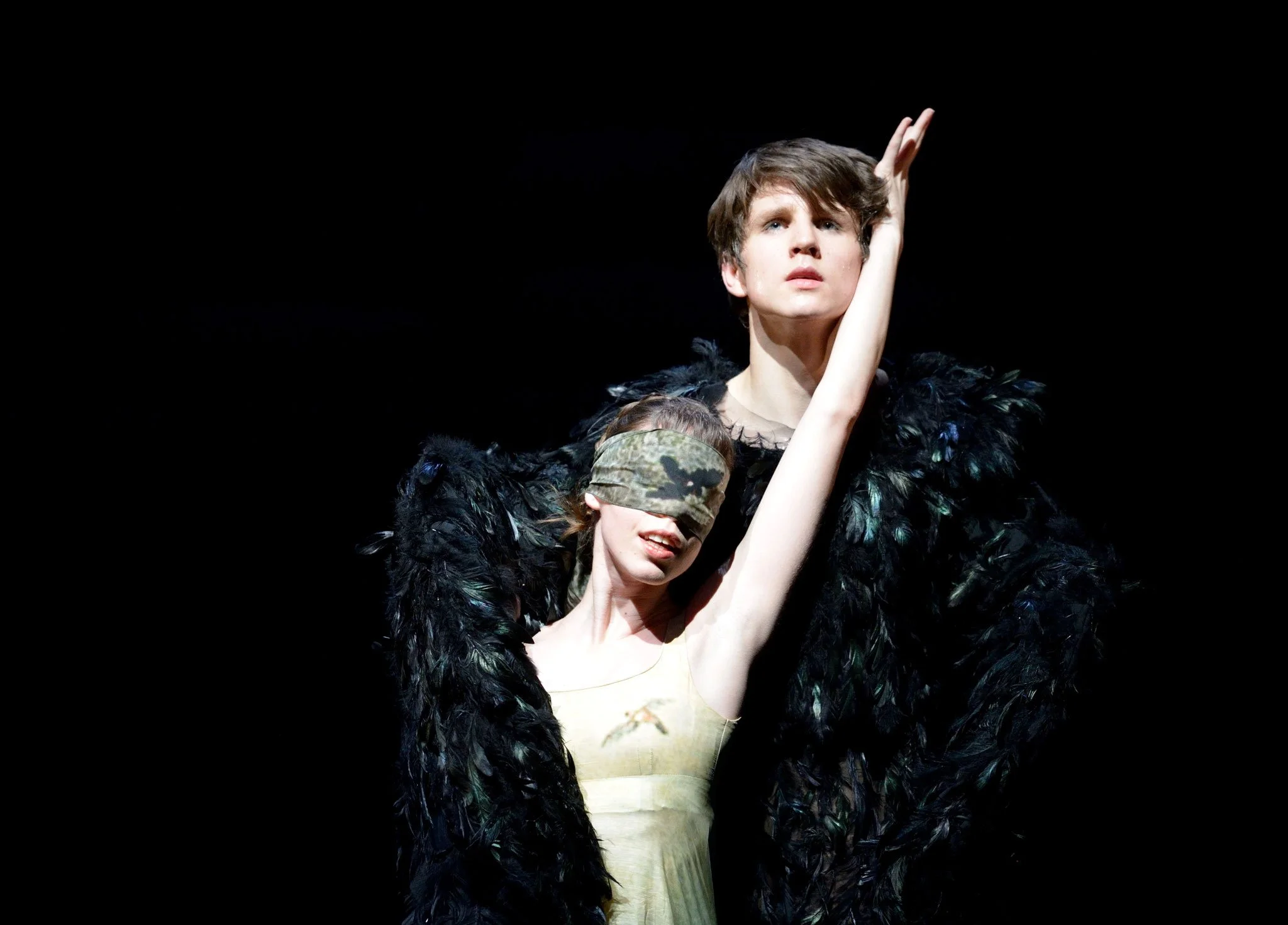 Two performers on stage, one with short hair and a feathered black costume, the other with light brown hair and a blindfold, performing with expressive gestures against a dark background.