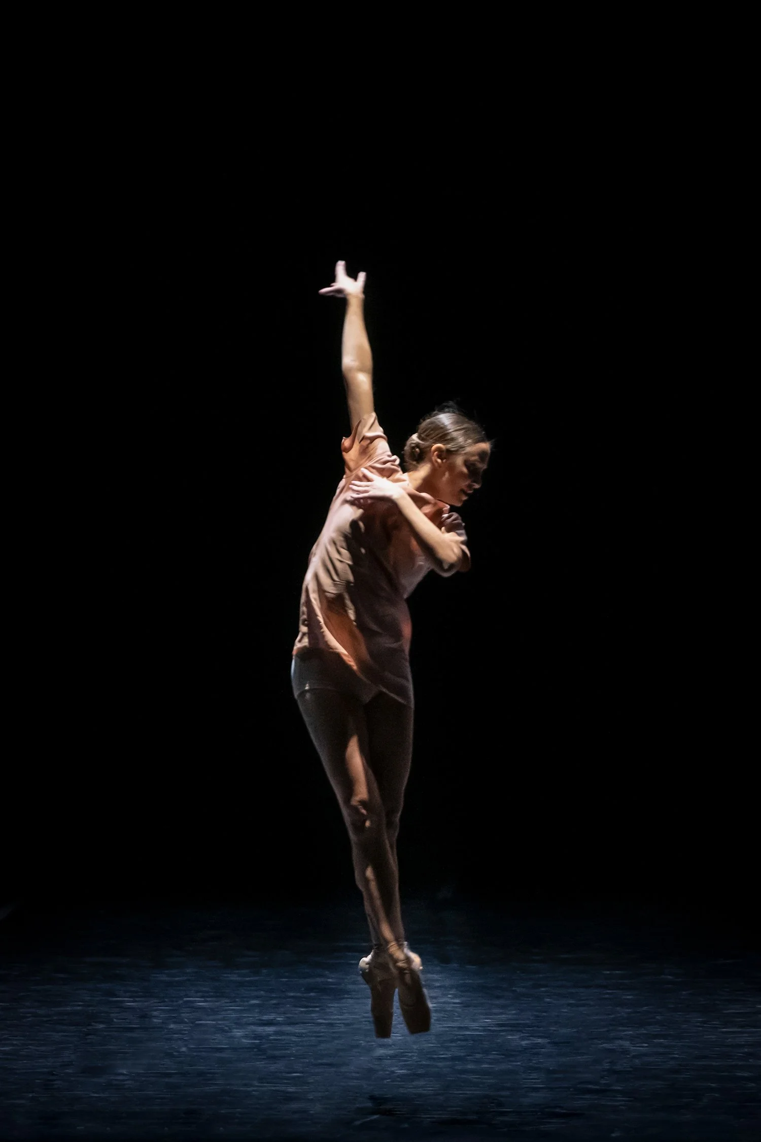 A female ballet dancer in a dance pose on stage with a dark background. She is mid-move, with one arm extended upward and the other across her chest, wearing a light-colored top and dark leggings.
