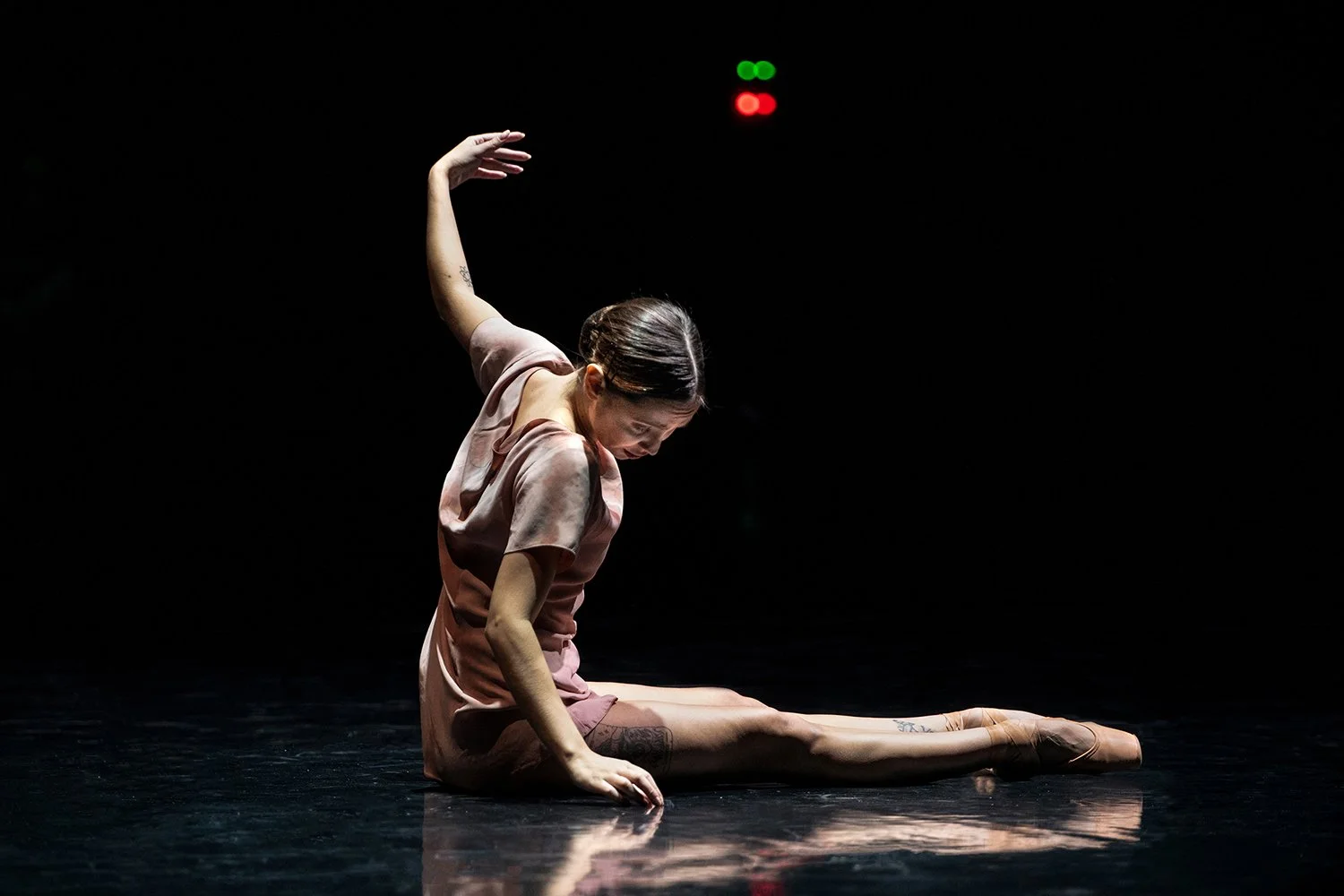 A female dancer in a pink dress performing a split on a dark stage, with her head bowed and arm raised. There is a red and green light in the background.