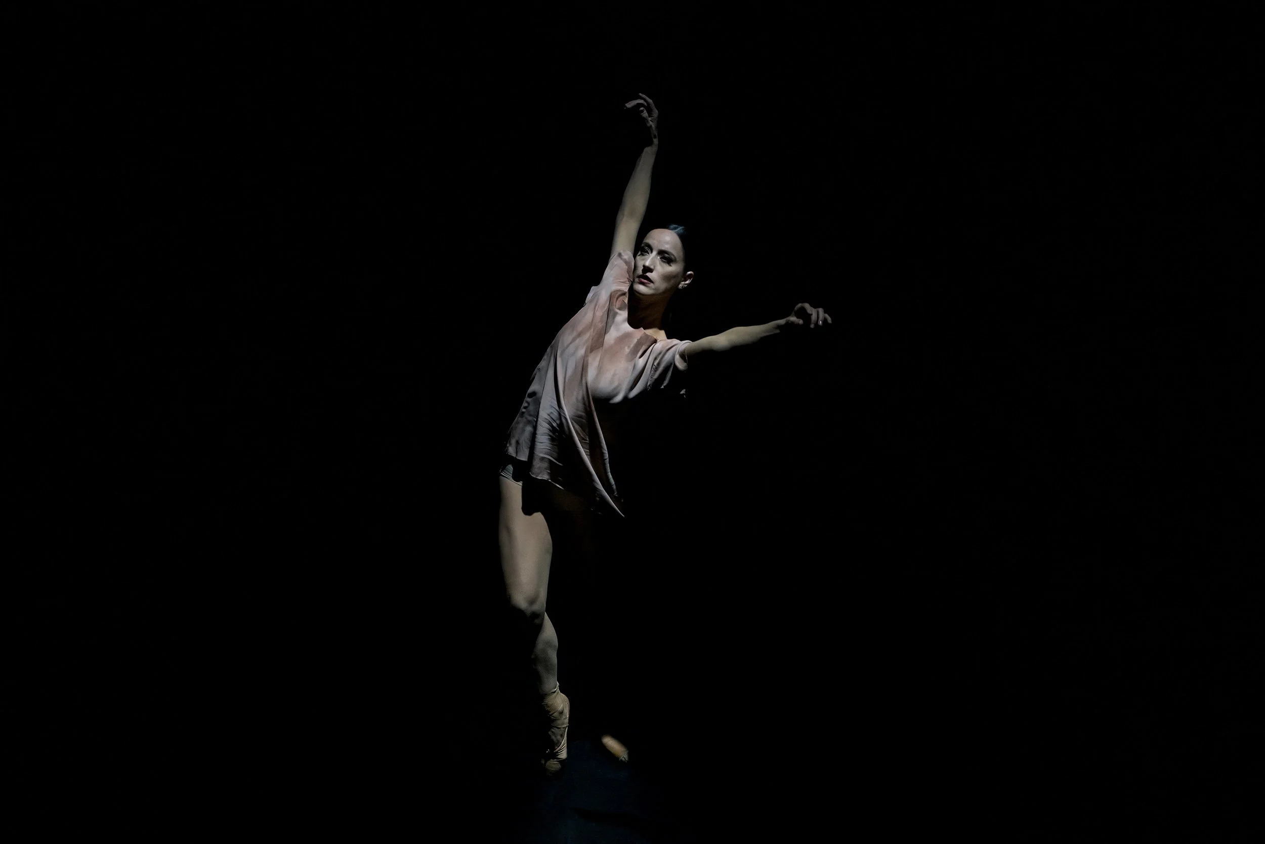 A female ballet dancer performing a pose on a dark stage, with one arm raised above her head and the other extended to the side.