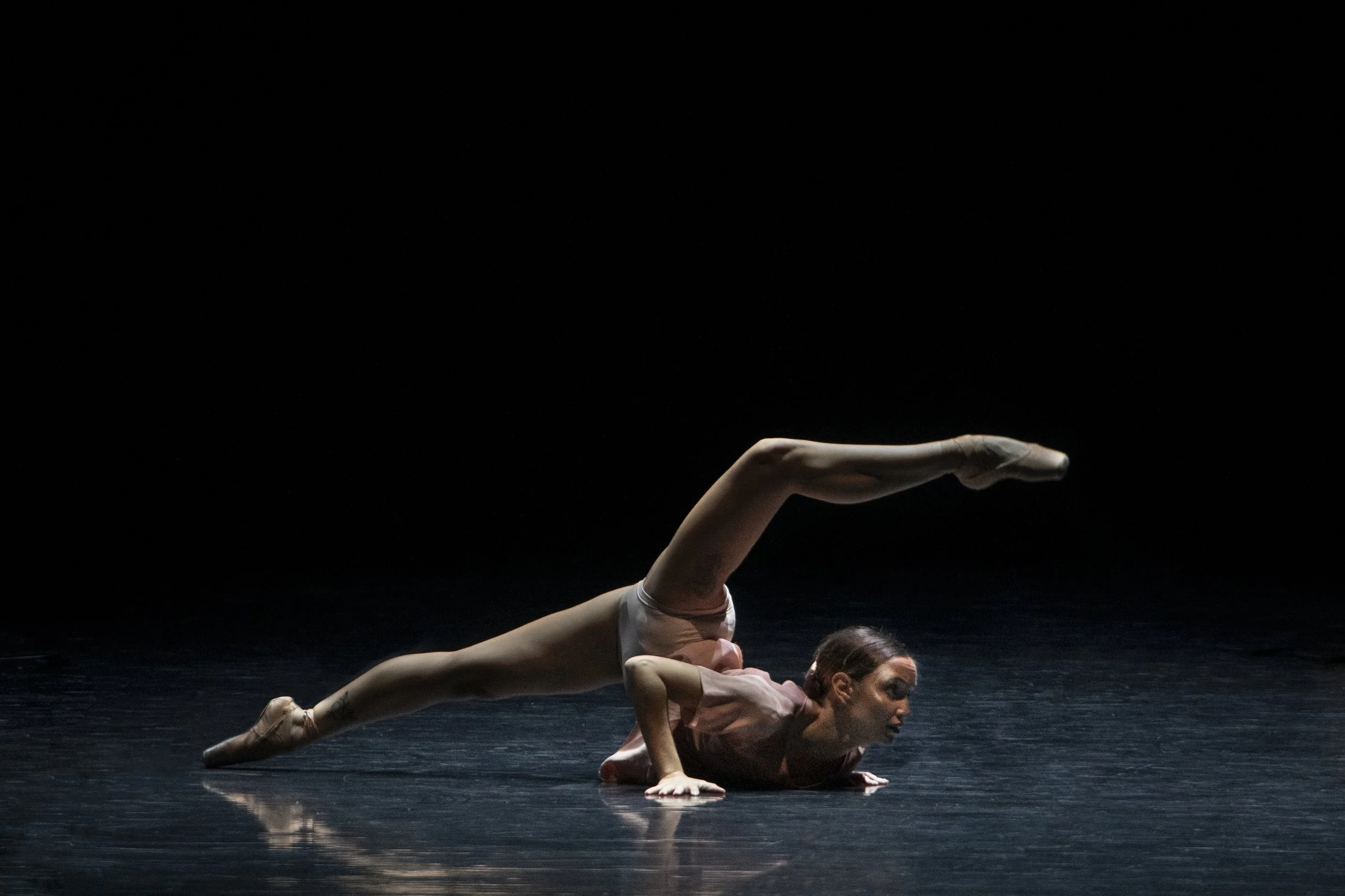 A ballet dancer is balancing on her hands with her legs extended in a split position in a dark studio.