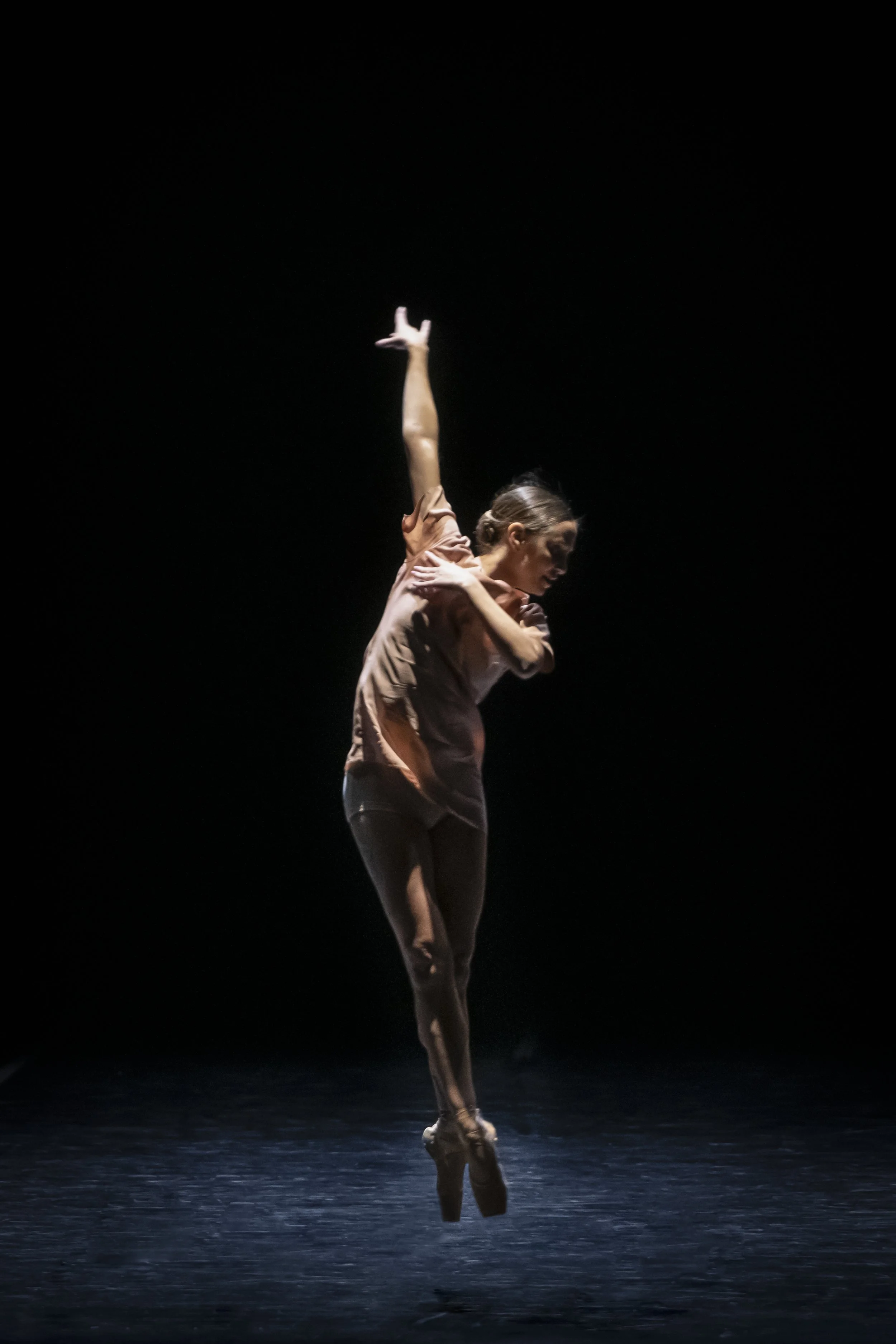 A female ballet dancer in a dance pose on stage with a dark background. She is mid-move, with one arm extended upward and the other across her chest, wearing a light-colored top and dark leggings.