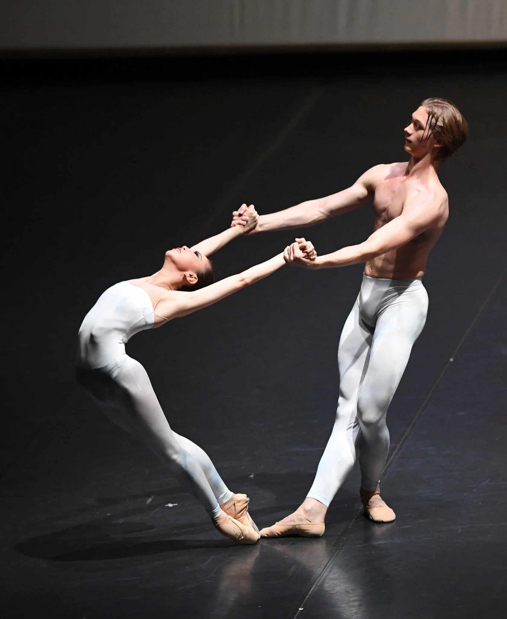 Two ballet dancers performing on stage, one standing and the other bent backwards, with their hands clasped.