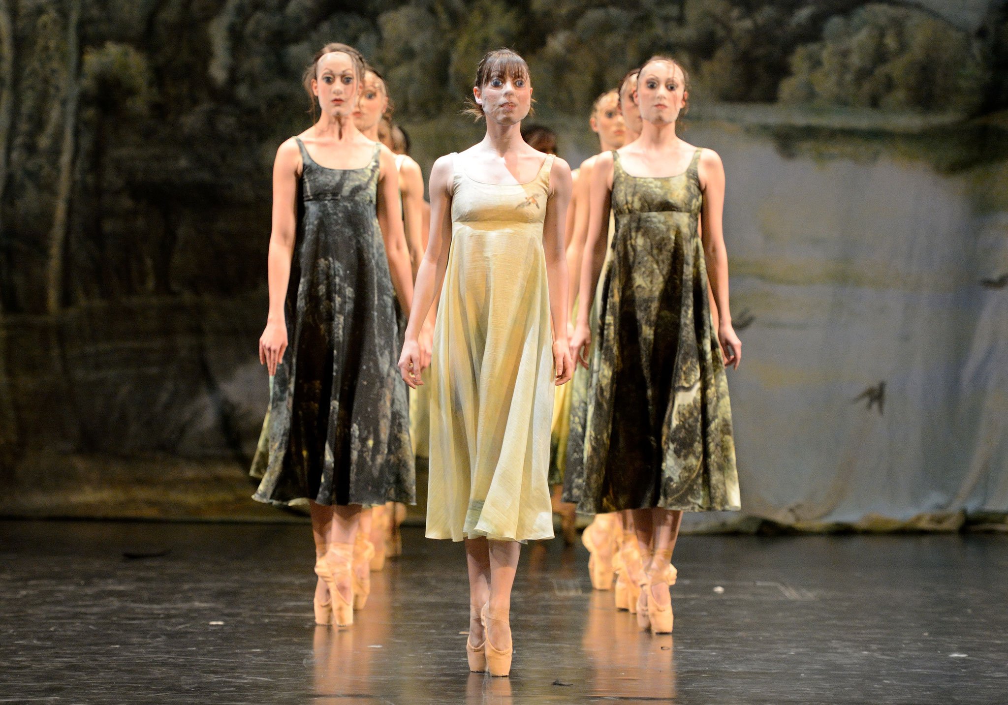 Group of female dancers performing on stage in flowing dresses and high heels, standing in a formation with forest backdrop.