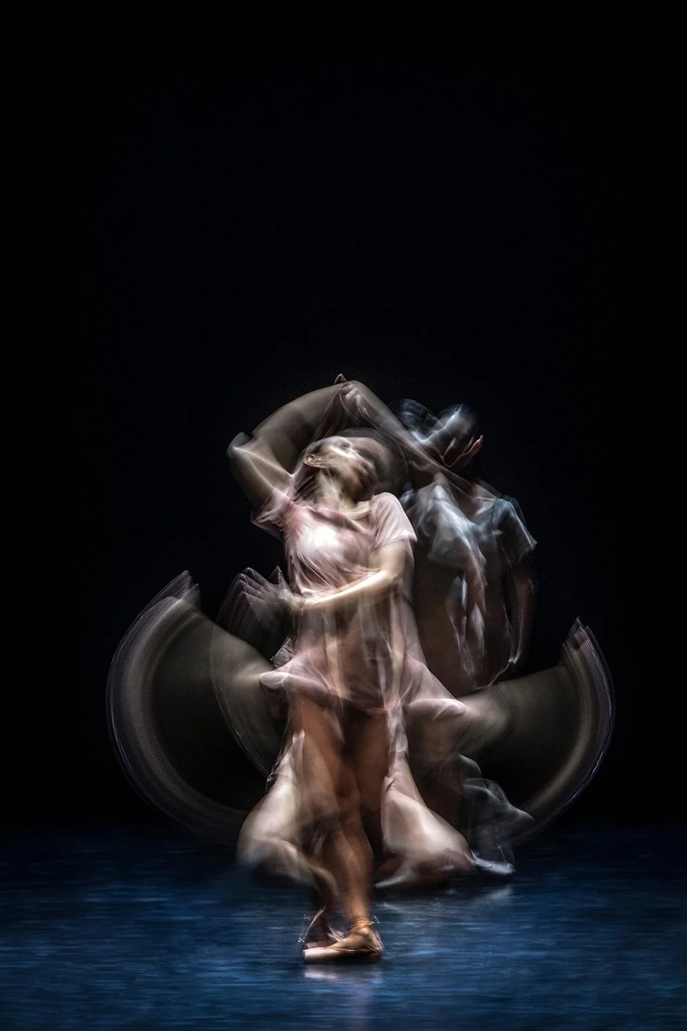 A dancer in a light-colored dress performs a movement on stage, with a dark background and motion blur creating a dynamic and abstract effect.