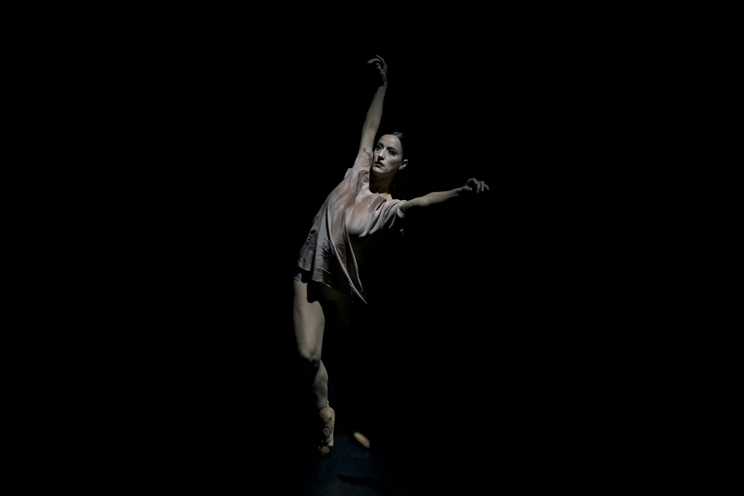A female ballet dancer performing a pose on a dark stage, with one arm raised above her head and the other extended to the side.