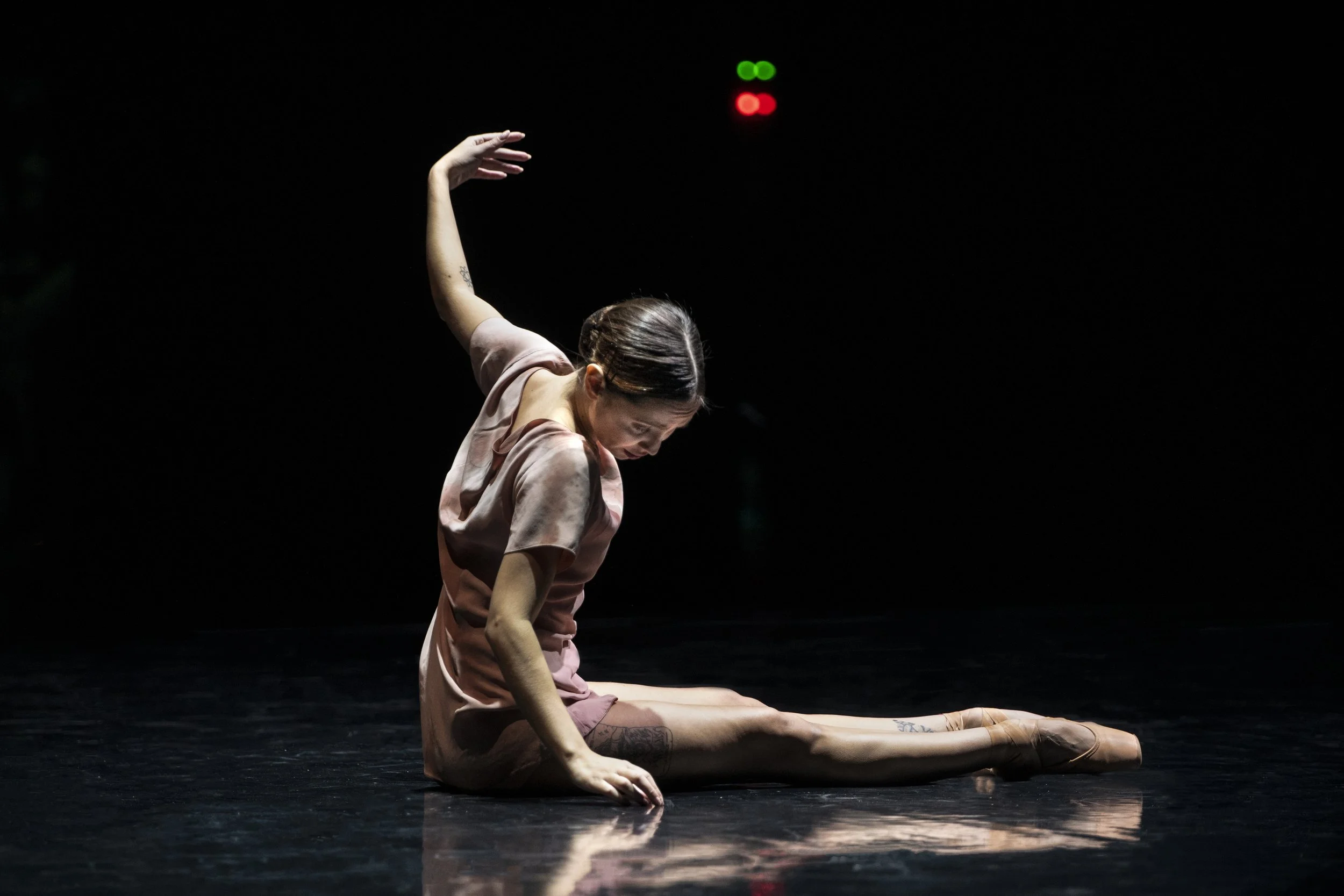 A female dancer in a pink dress performing a split on a dark stage, with her head bowed and arm raised. There is a red and green light in the background.