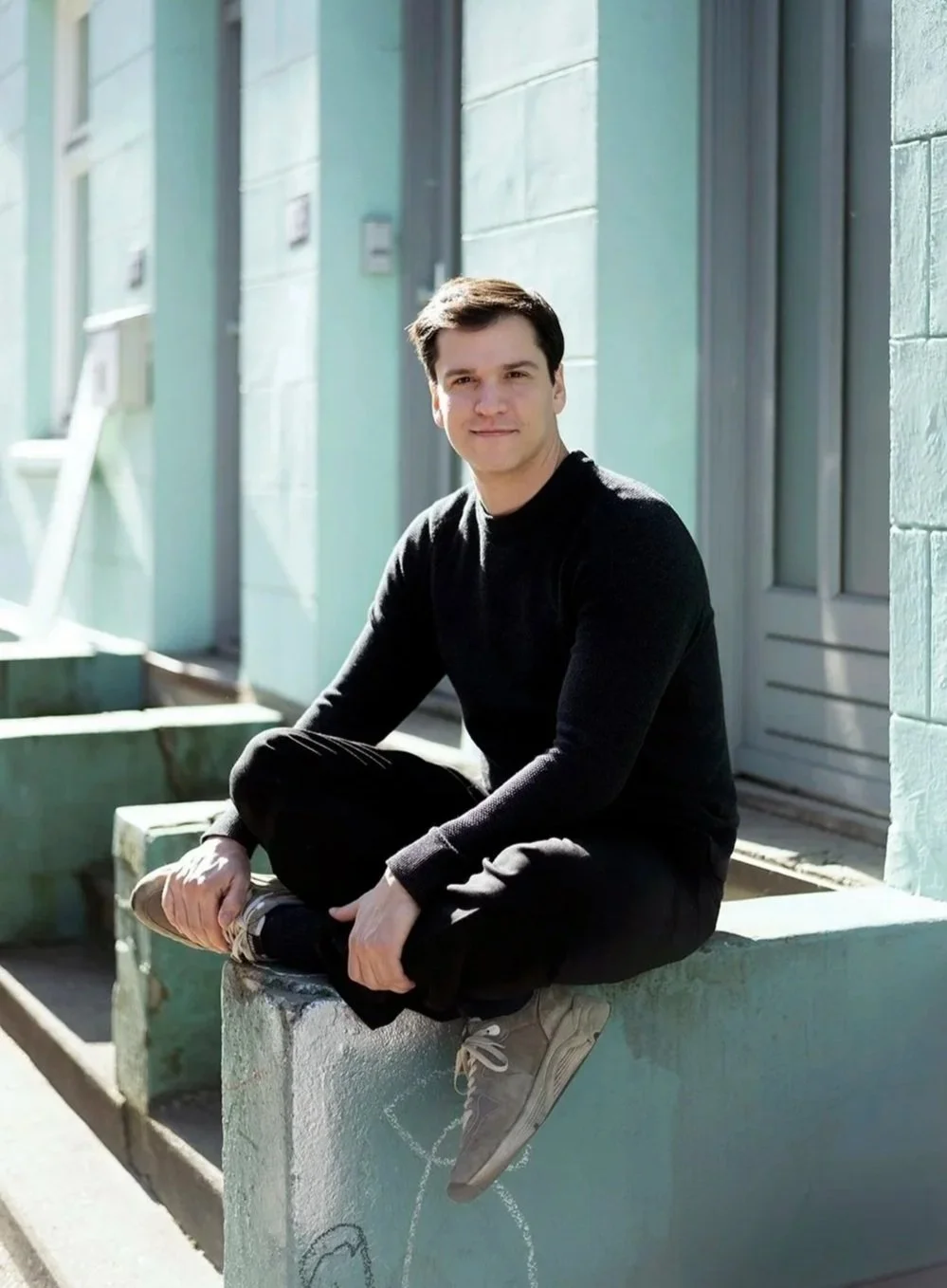 Portrait of choreographer Demis Volpi in black clothing, seated on a ledge outside a building and smiling at the camera.
