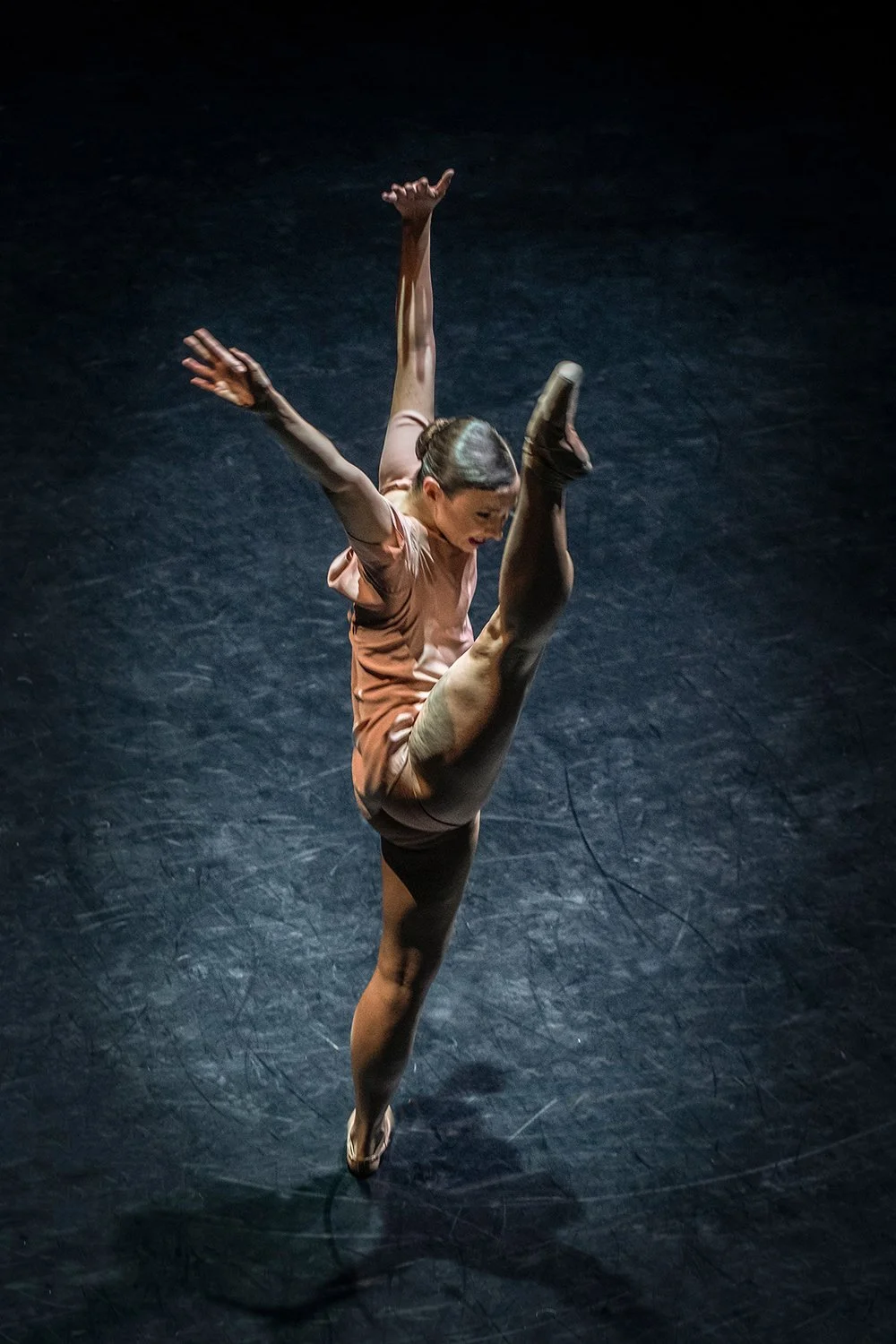 A female ballet dancer in a peach costume performs a high leg extension with her arms extended outward, on a dark stage.