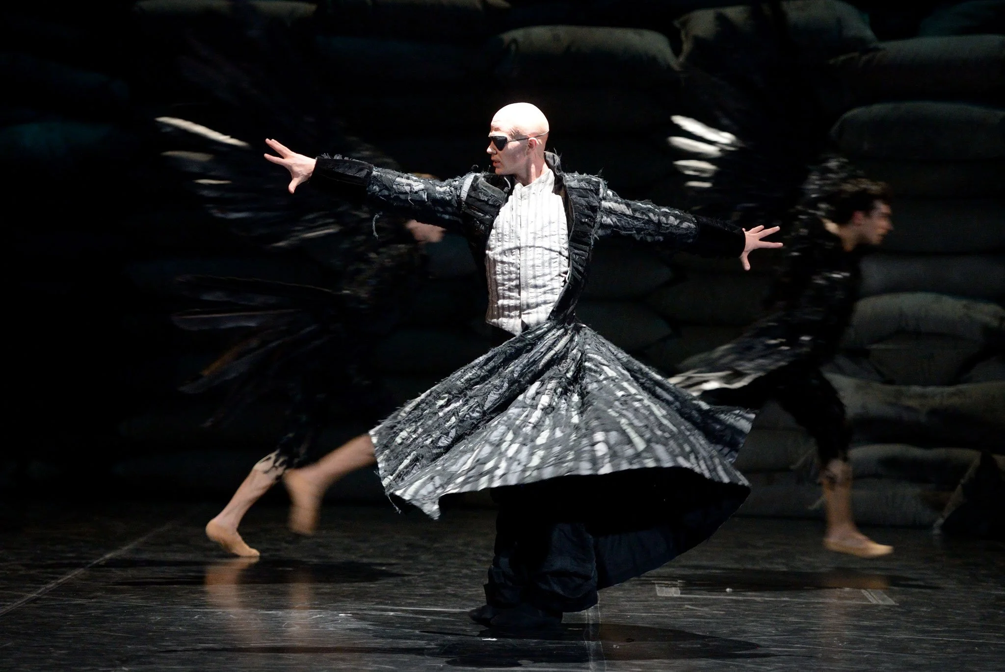 Dancer with outstretched arms in costume, on stage with reflections on the black floor.