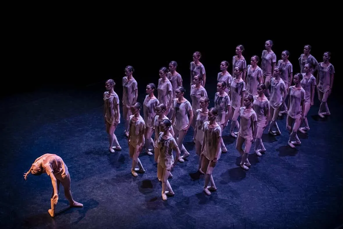 A group of young dancers performing on stage, with one dancer bending backwards and extending her arms, while the others stand in formation behind her, all dressed in matching costumes under stage lighting.