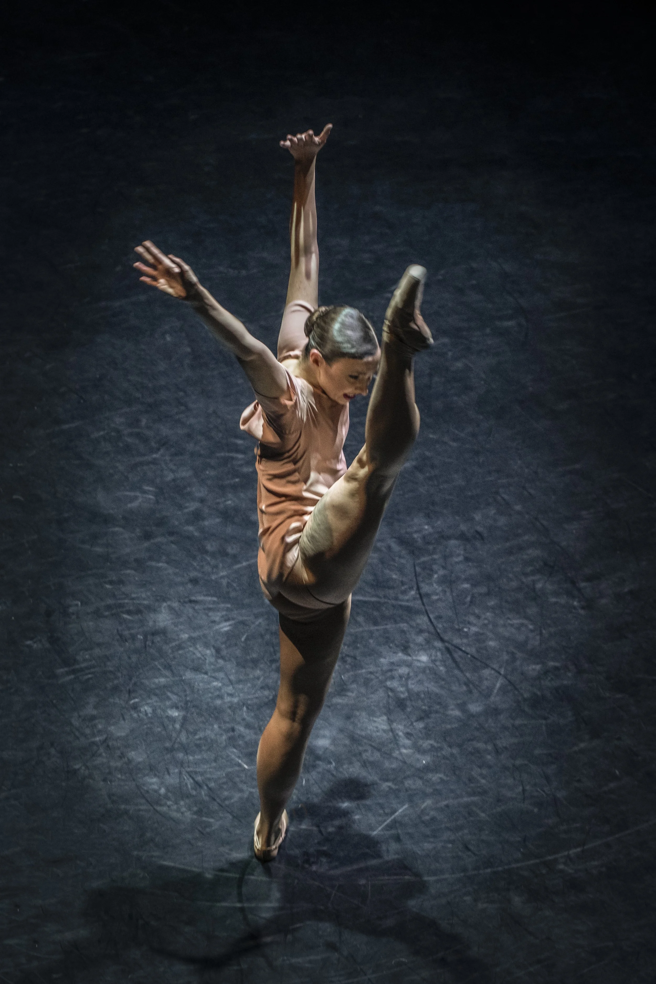 A female ballet dancer in a peach costume performs a high leg extension with her arms extended outward, on a dark stage.