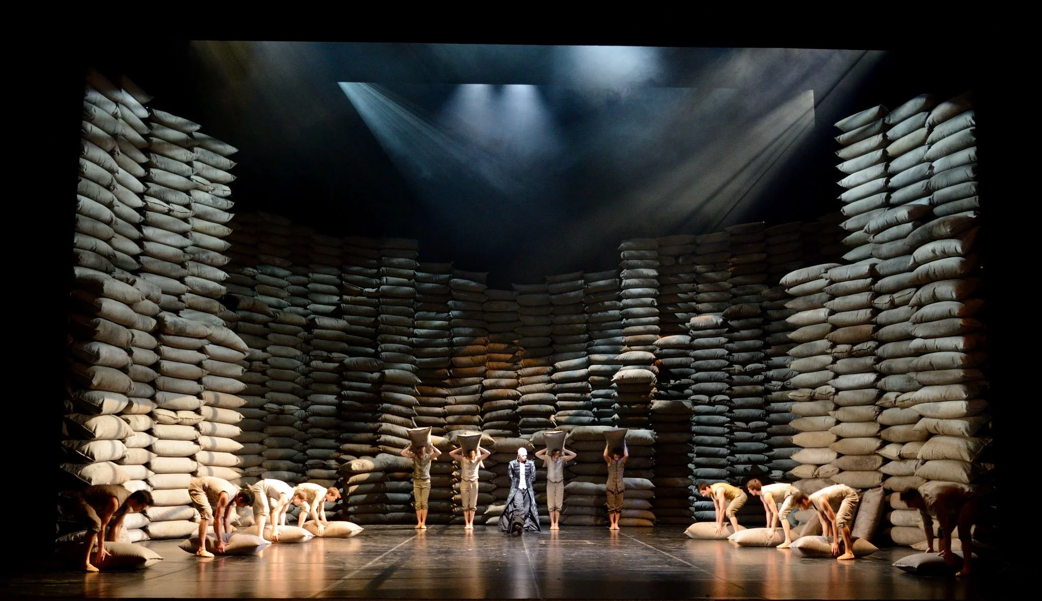 The image shows a theatrical stage set resembling an indoor arena or storage warehouse with tall, stacked sandbags forming walls and levels. People are performing, some carrying large bags on their heads, and others are kneeling or bowing on the stag