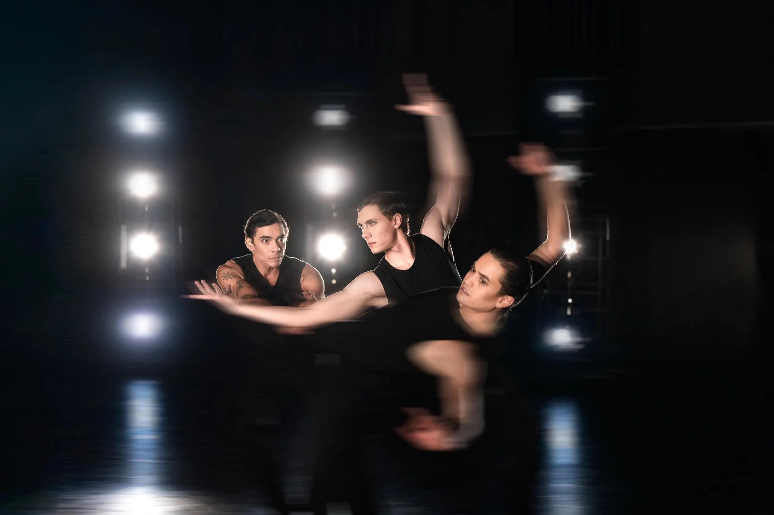 Three ballet dancers performing on stage with bright lights behind them, captured mid-movement.