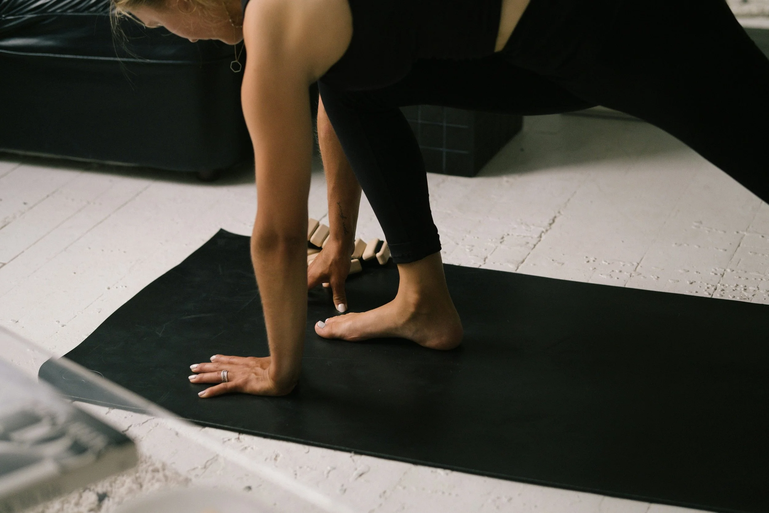 A person practicing yoga or stretching on a black yoga mat, in a room with a white textured floor, wearing black leggings and a sleeveless top.