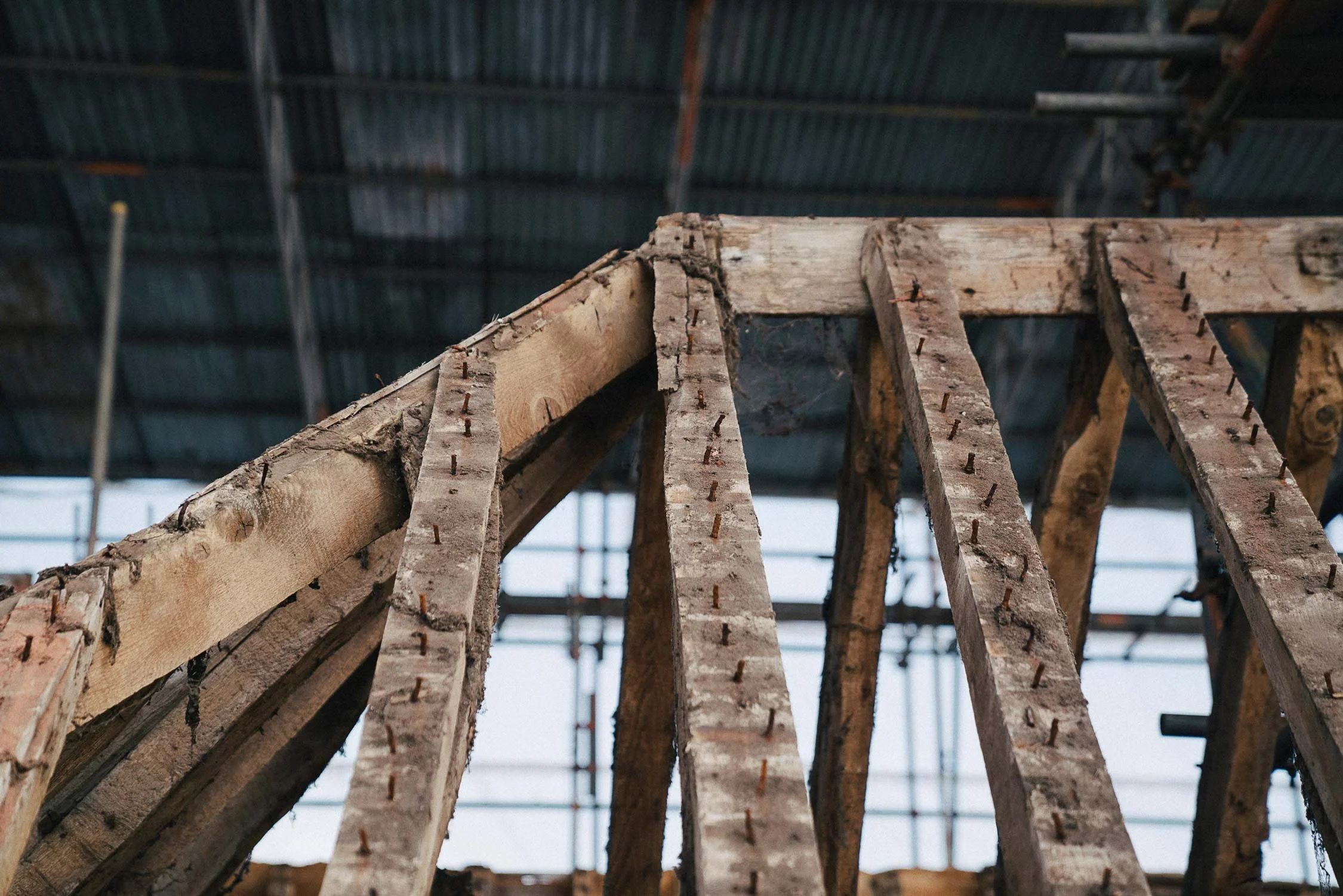 Close-up view of a wooden roof structure under construction, showing wooden beams and nails, with a metal roof in the background.