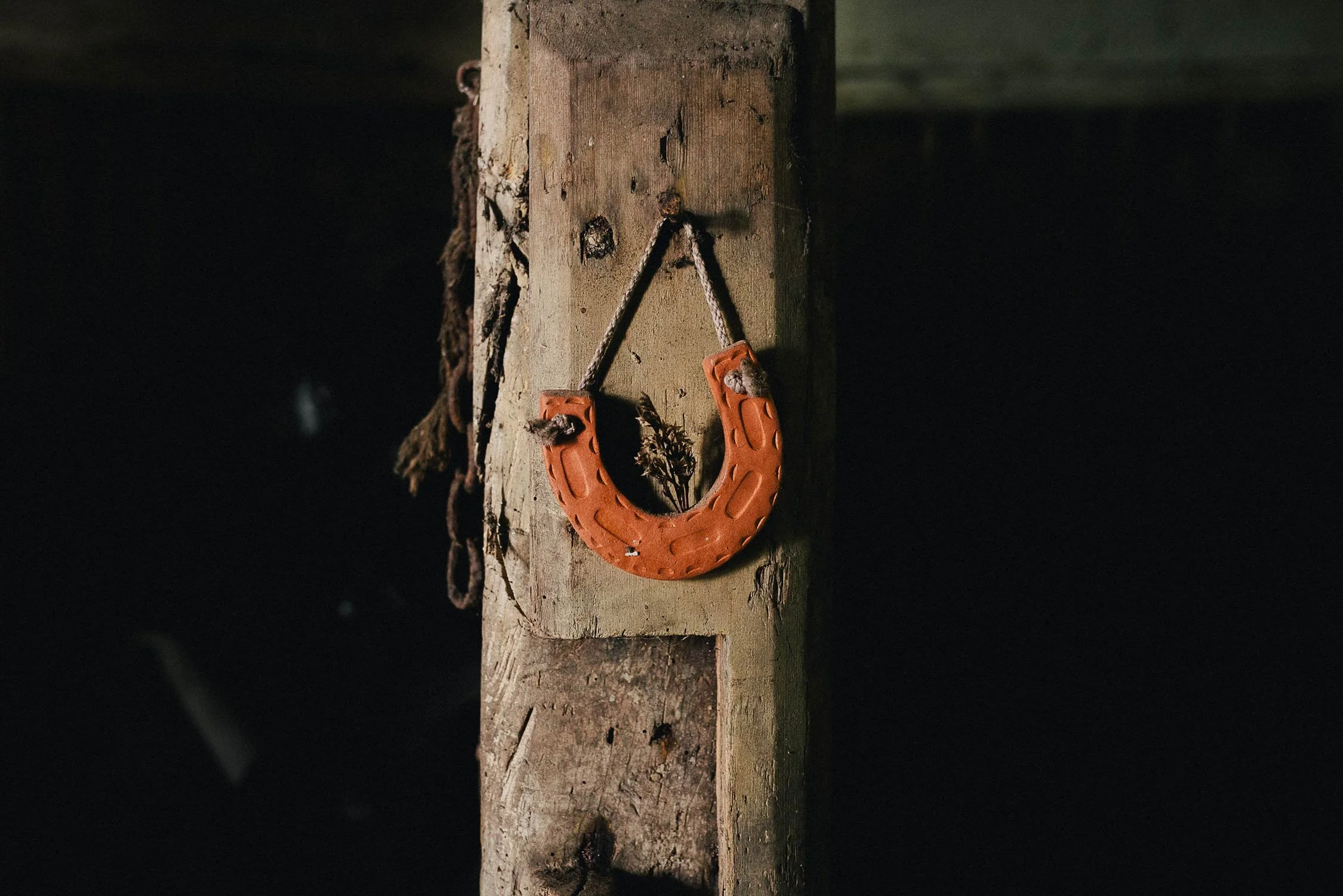 Close-up of a weathered wooden beam with a small, orange horseshoe hanging on it, attached by a rope, with a dried flower underneath.