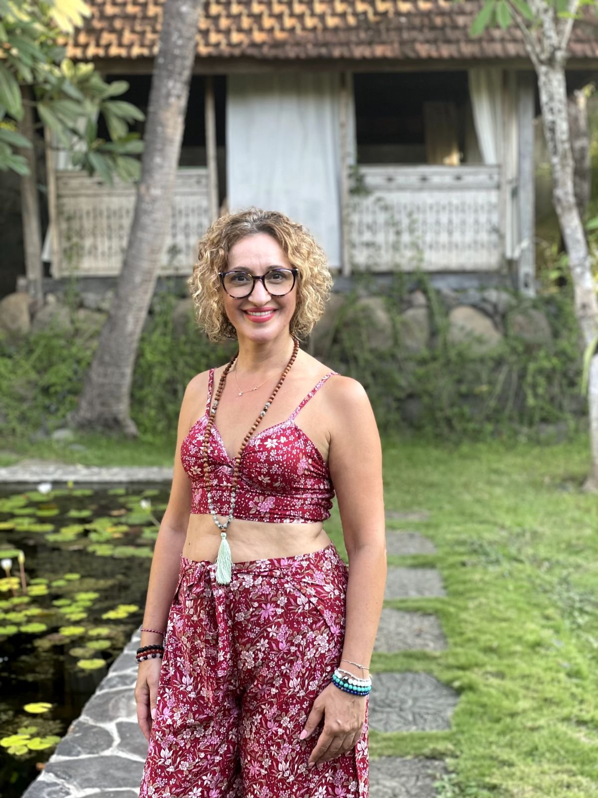 A woman with light curly hair and glasses smiling outdoors in a red floral crop top and matching flowy pants, surrounded by green grass, trees, a pond with lily pads, and a wooden house with a tiled roof in the background.