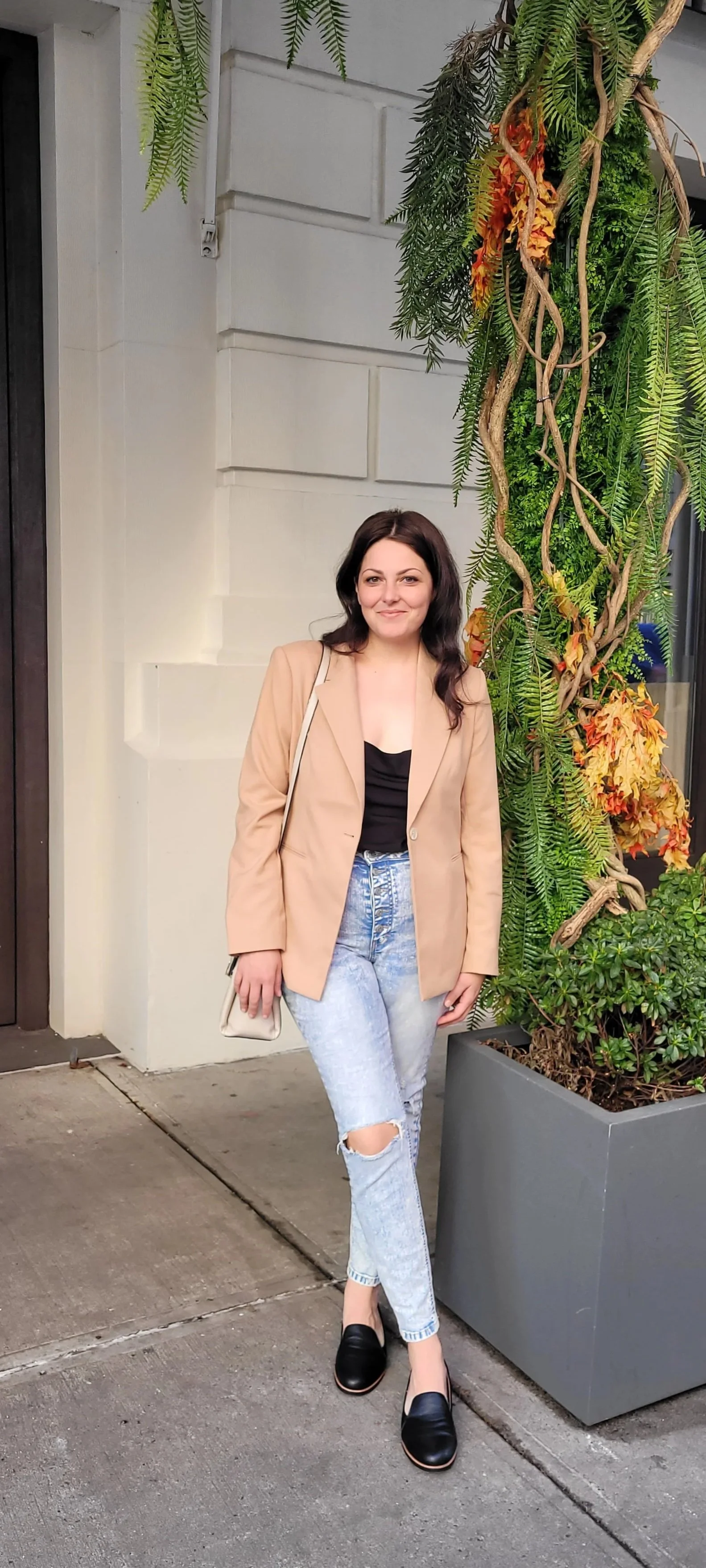 A young woman with turned black hair standing next to a large green and orange plant in an outdoor urban setting, wearing a beige blazer, black top, light blue ripped jeans, and black slip-on shoes.