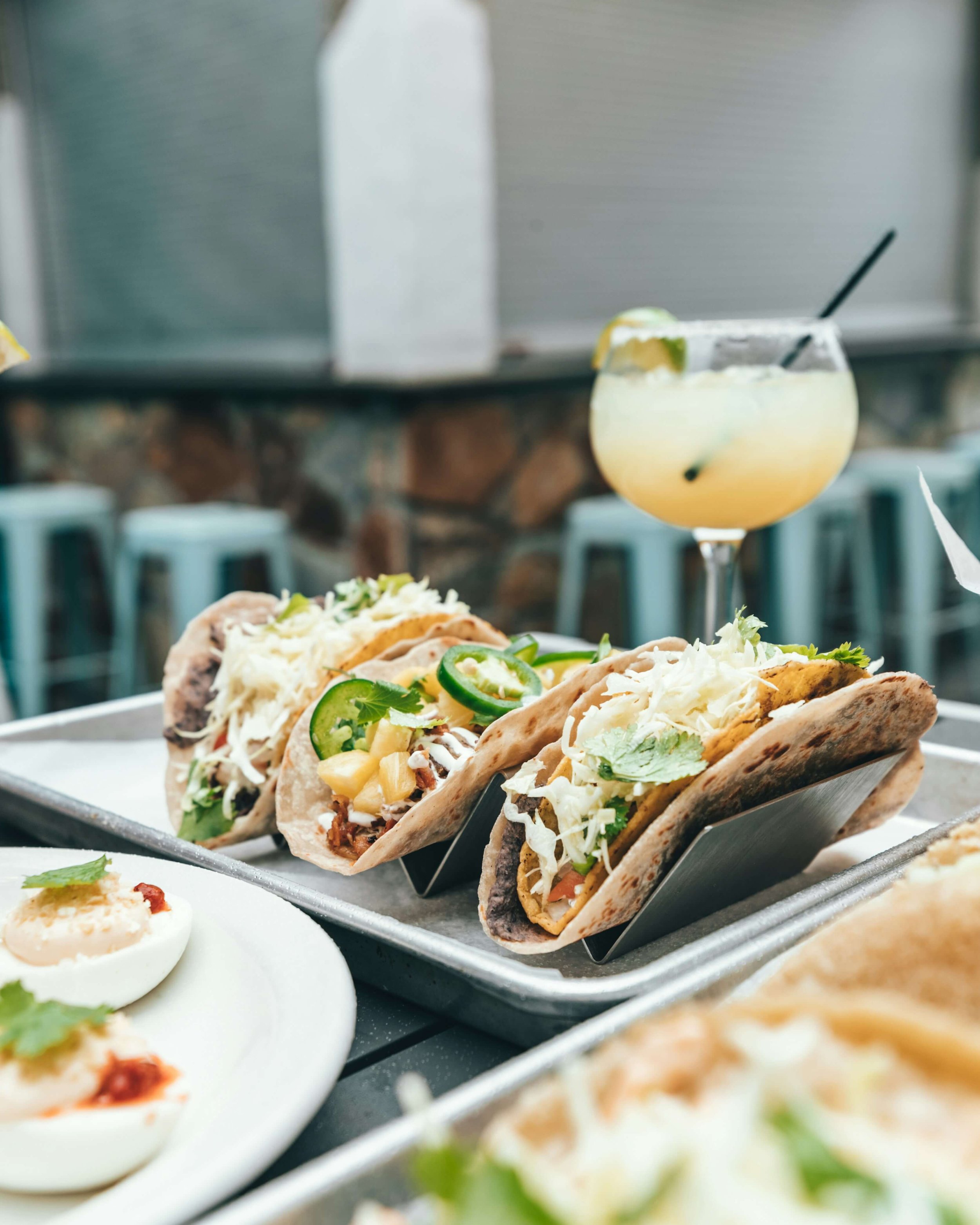 Tray of tacos with various fillings including jalapeños and cilantro, a cocktail glass with a lime garnish, and a plate of deviled eggs on an outdoor table with bar stools in the background.