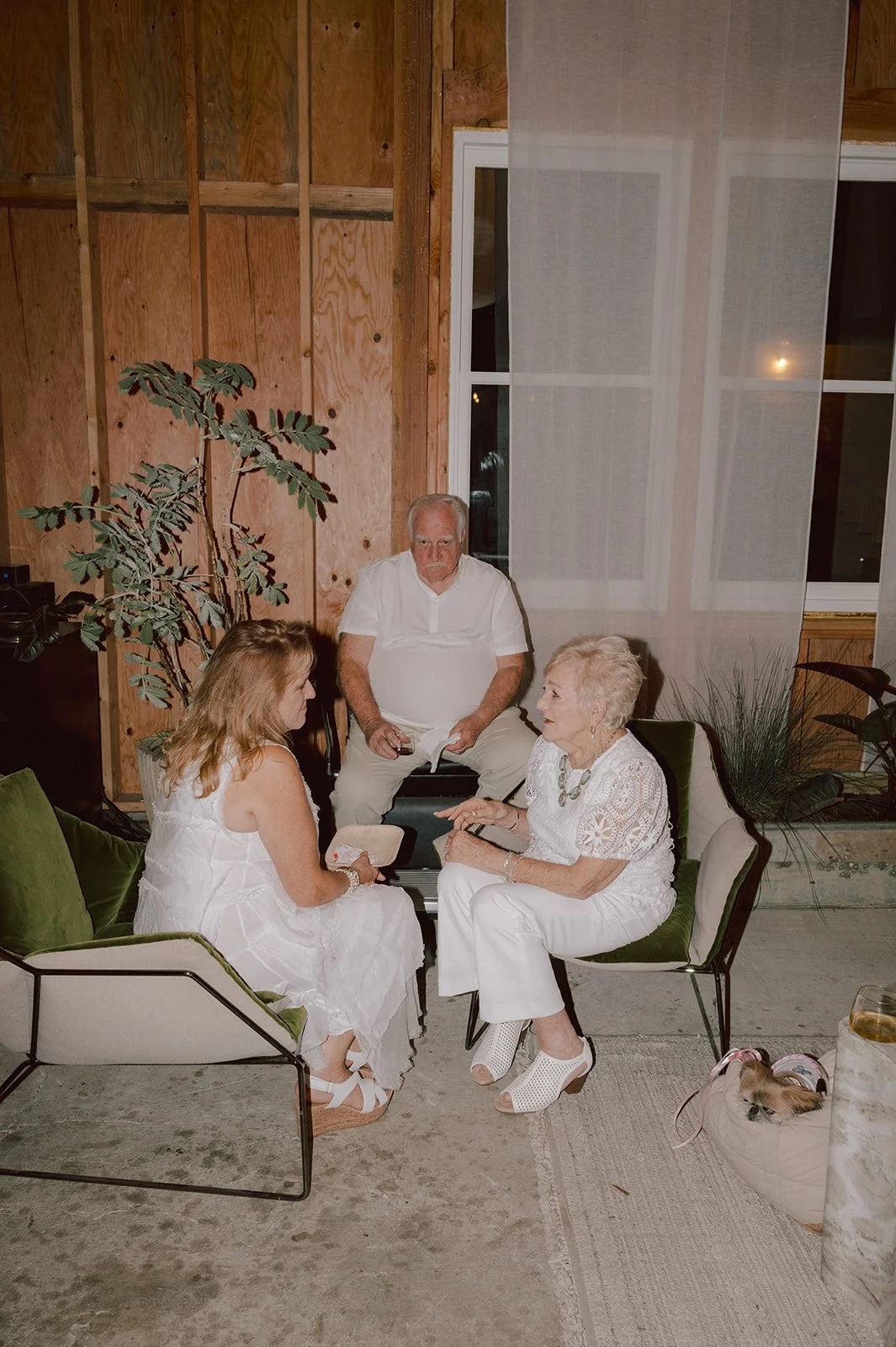 Four people, two women and two men, are sitting and chatting inside a rustic room with wooden walls. One woman has white hair, wears a white outfit and is sitting in a green chair. The younger woman wears a white dress and sandals, and is seated on a
