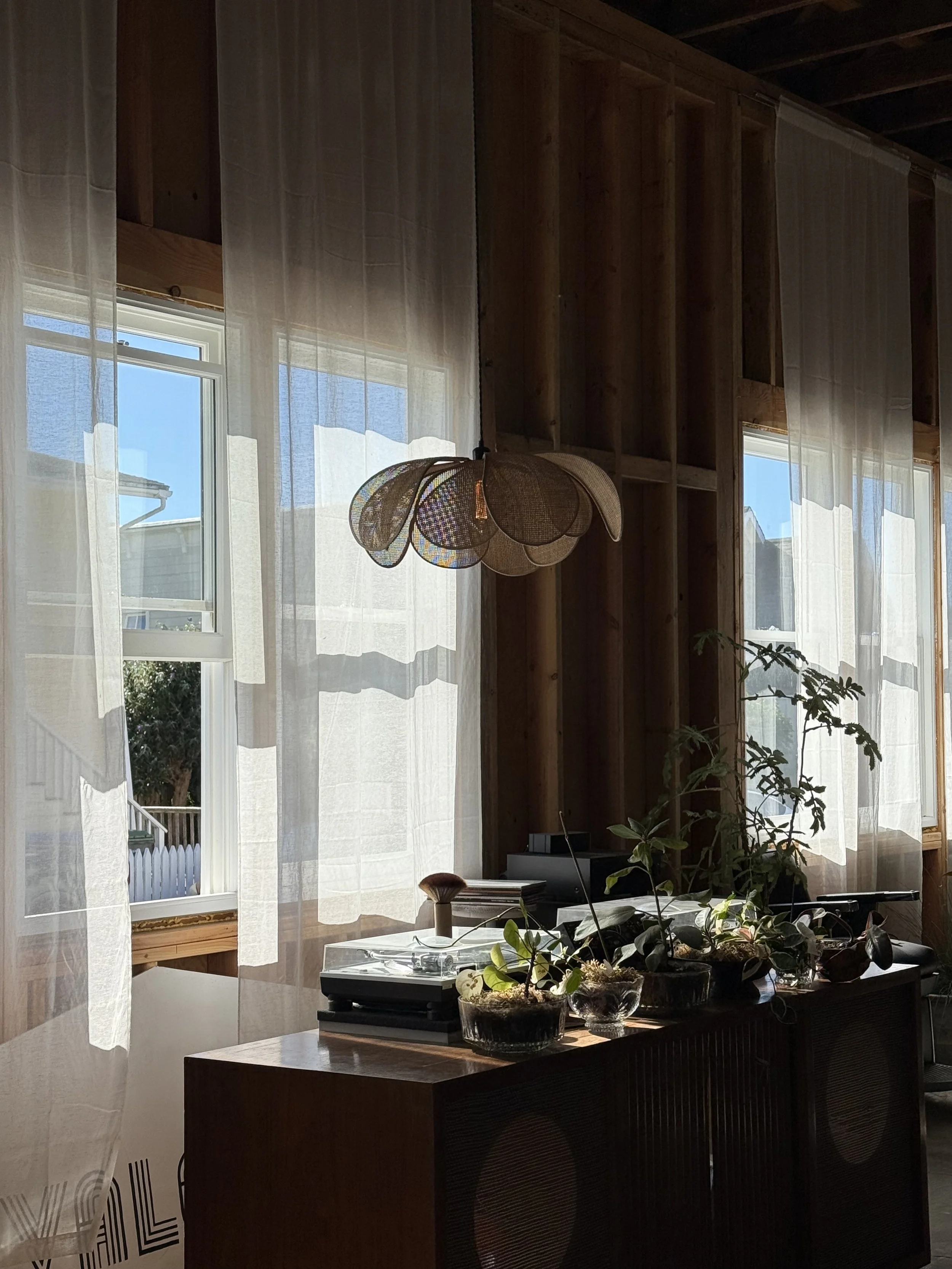Interior view of a room with wooden walls, sheer white curtains, a modern hanging light fixture, a potted plant, and a table with a record player and other objects.