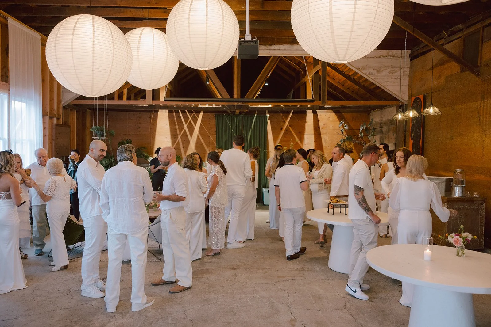 People dressed primarily in white attire mingling at a social gathering or celebration inside a rustic wooden venue with large round paper lantern lights hanging from the ceiling.