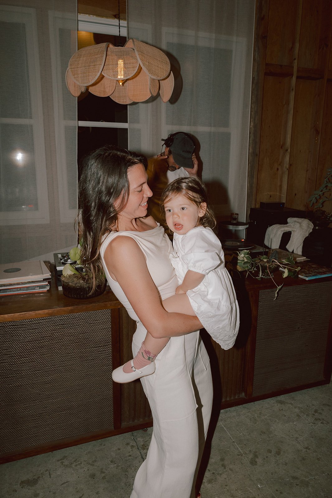 A woman in white holding a young girl in a white dress indoors at night, with a wooden cabinet and a person wearing a cap in the background.