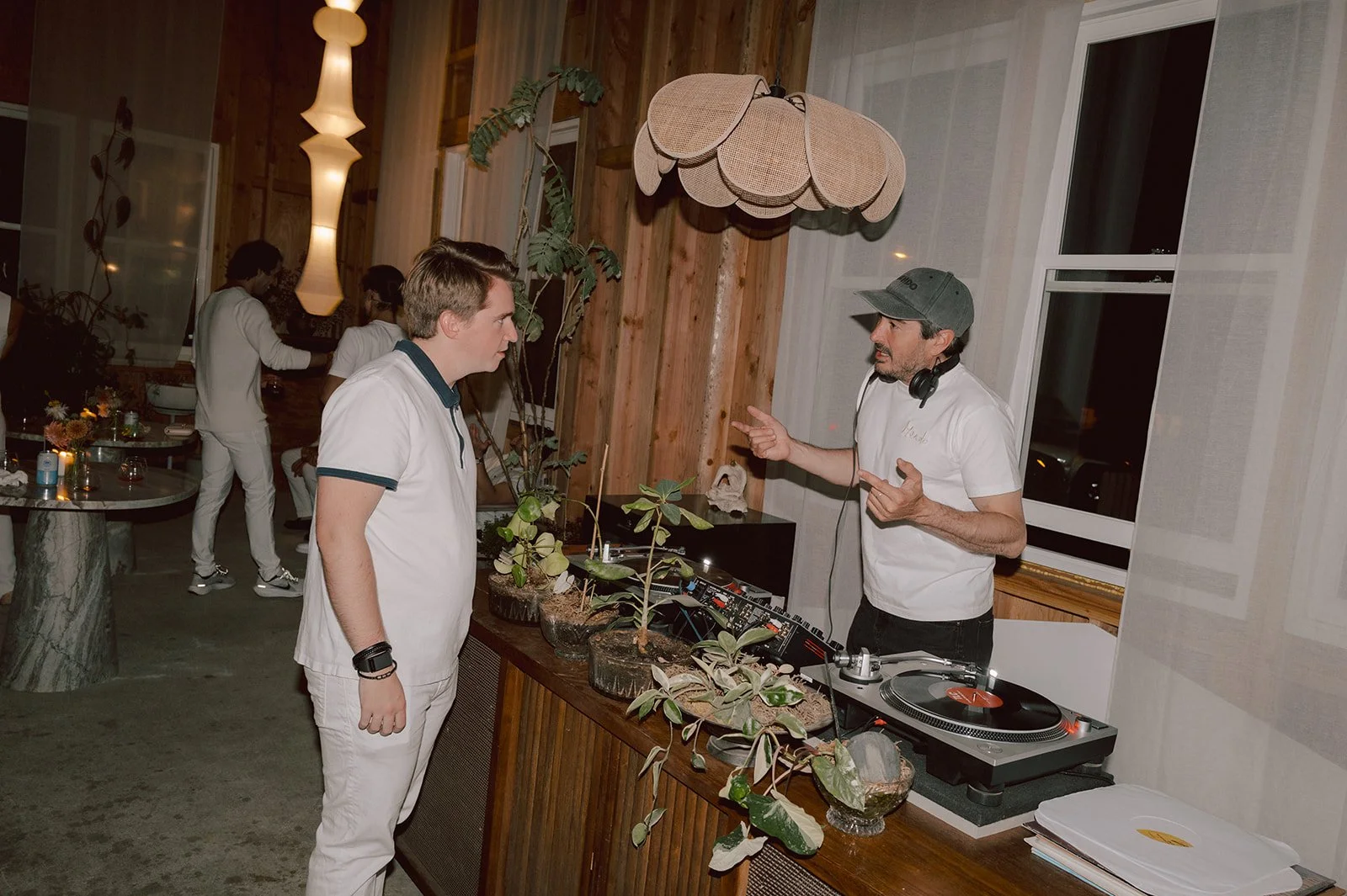 A DJ wearing a gray cap and white t-shirt is speaking to a man in a white polo shirt, surrounded by potted plants on a wooden counter, inside a warmly lit room.