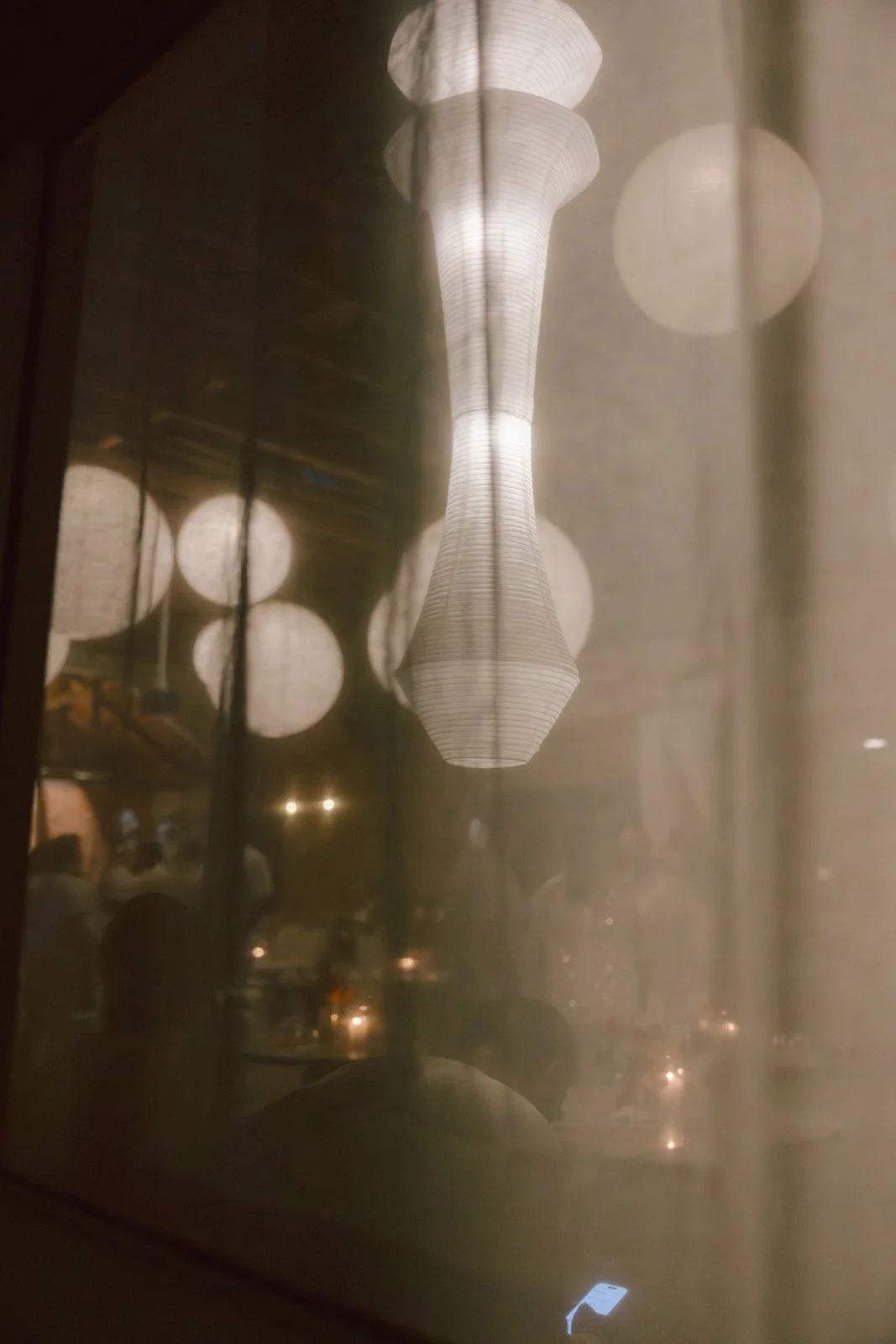 A view through a drapped window in our dimly lit space, showing decorative hanging lanterns and faint silhouettes of people dining.