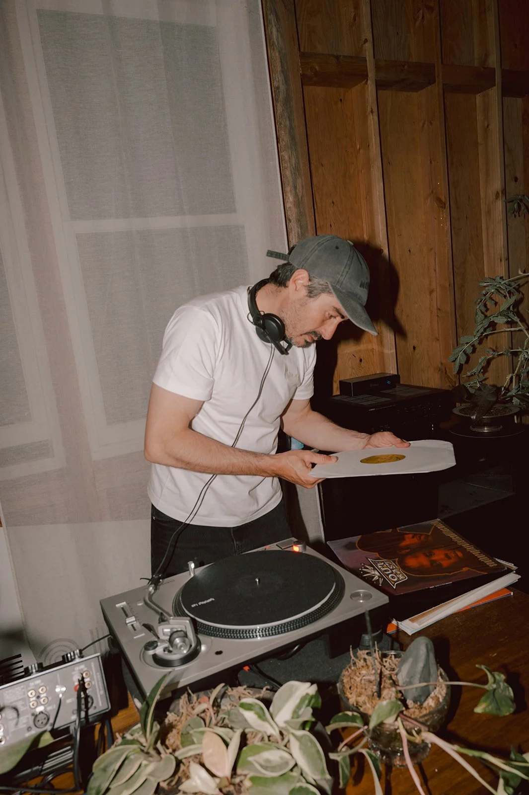 A man wearing a gray baseball cap, white t-shirt, and headphones leaning over a vinyl record at a DJ setup, with a turntable, vinyl records, and plants on the table, and a wooden wall in the background.