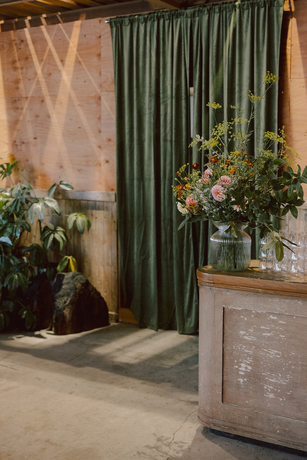 A rustic indoor setting with a wooden wall and green curtains, a plant in the corner, a large rock, and a wooden cabinet topped with a vase of mixed flowers and glasses.