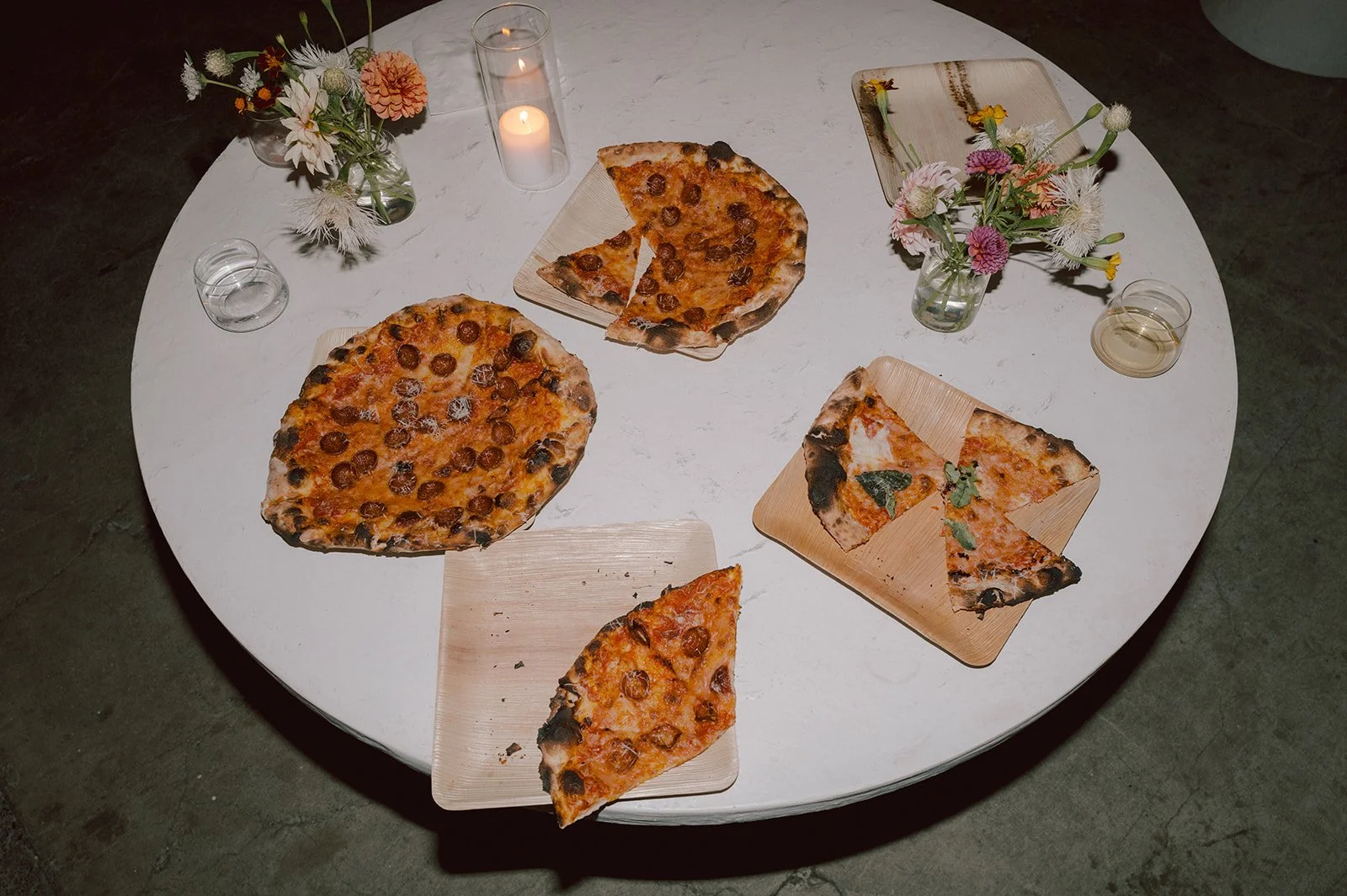 Round table with three slices of pizza, two whole pizzas, and floral centerpieces with candles.