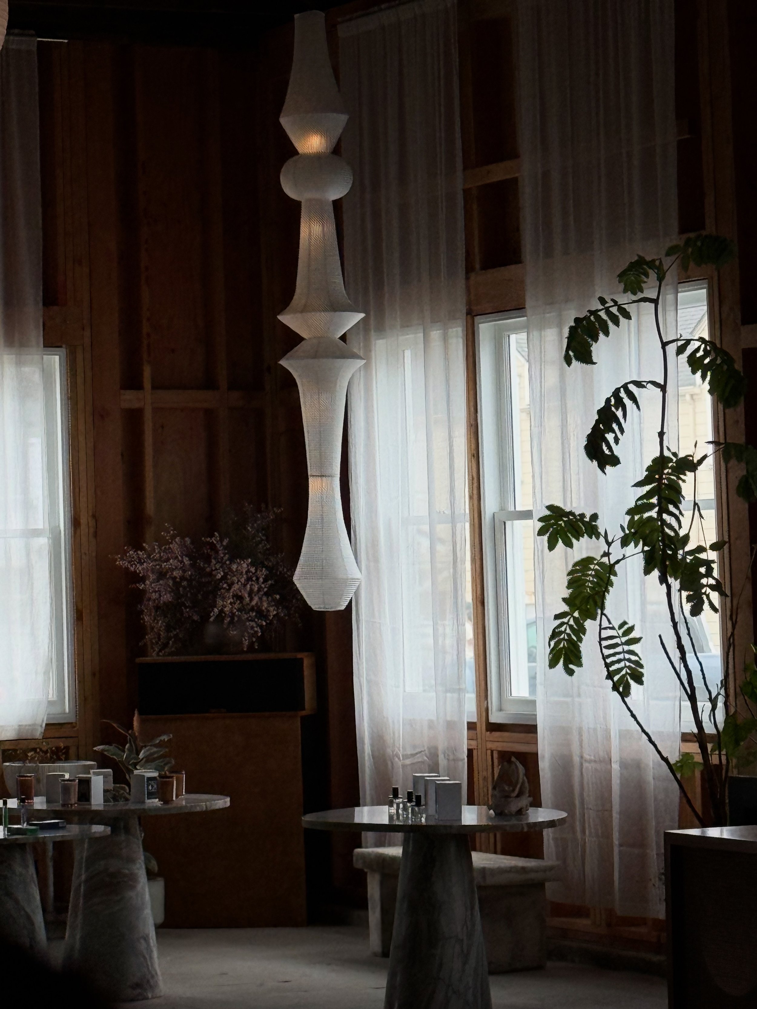 Interior of a room with wooden walls, large windows with sheer curtains, a tall, white, sculptural pendant light, a small table with chess pieces, and a leafy houseplant.