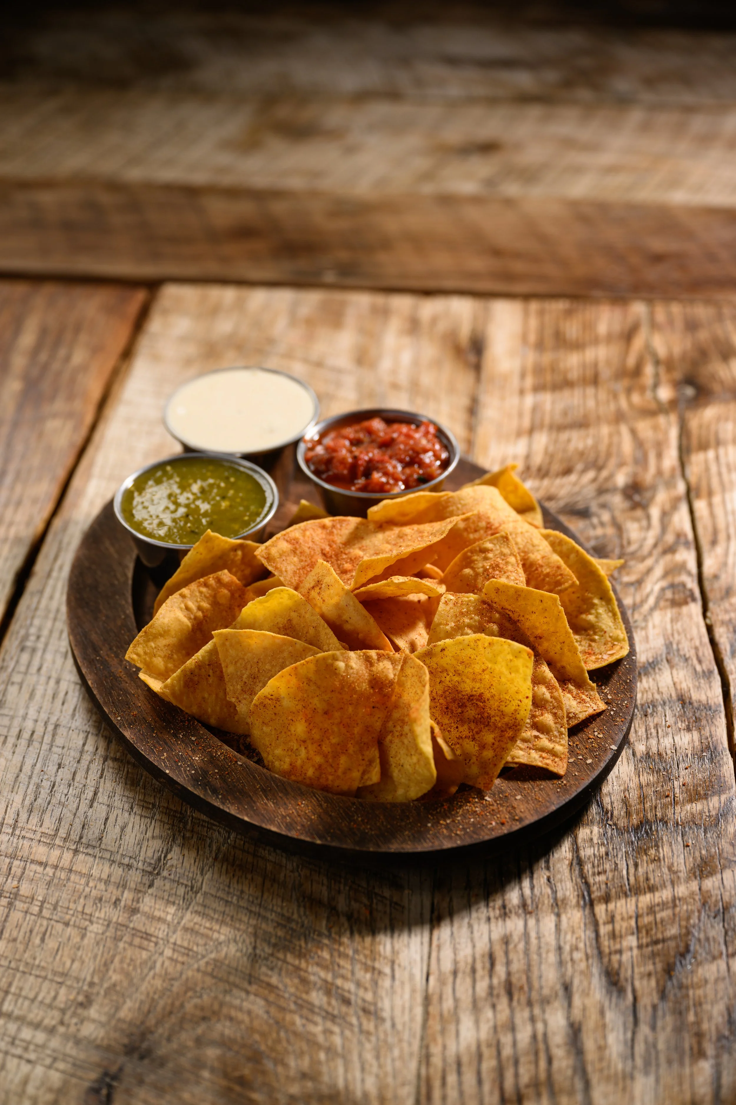 Plate of crispy tortilla chips with three small bowls of salsa, guacamole, and sour cream on a rustic wooden table.