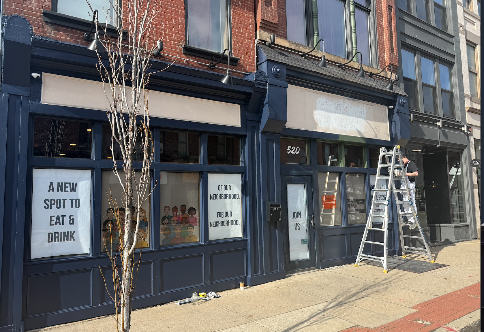 Construction workers are painting the exterior of a storefront with dark blue trim. Ladders and paint supplies are on the sidewalk. The windows display signs with messages and illustrations of diverse people.