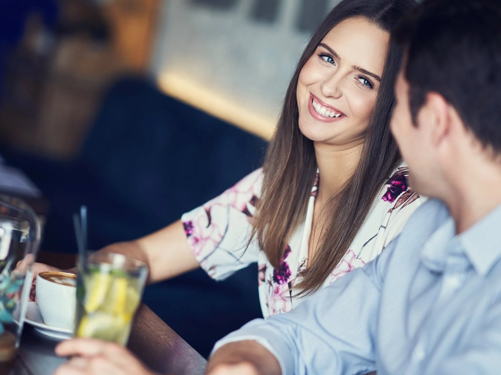 A woman smiling and talking to a man at a cafe table, with drinks and coffee in front of them.